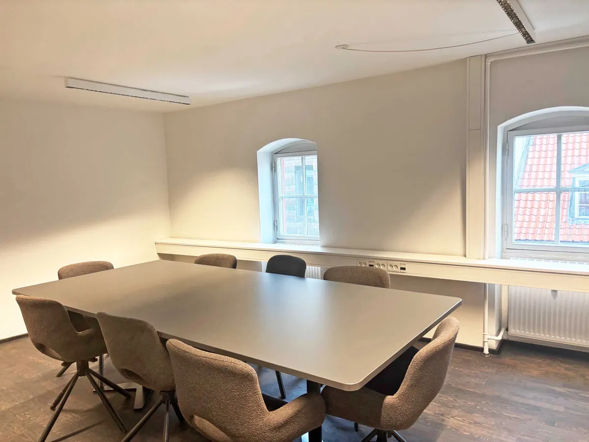 Moderne kontor med lyse trægulve og minimalistisk møblering. Et stort bord med flere stole, tæt på en slank radiator og to store vinduer med udsigt til røde tag.