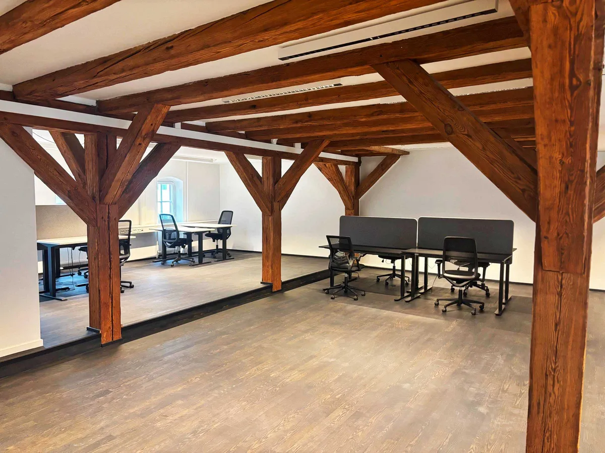 Værelse med lyse loftsbjælker og trægulv. To grupper med kontorbord og stole. Lyse vægge og vinduer. Modernt design.
