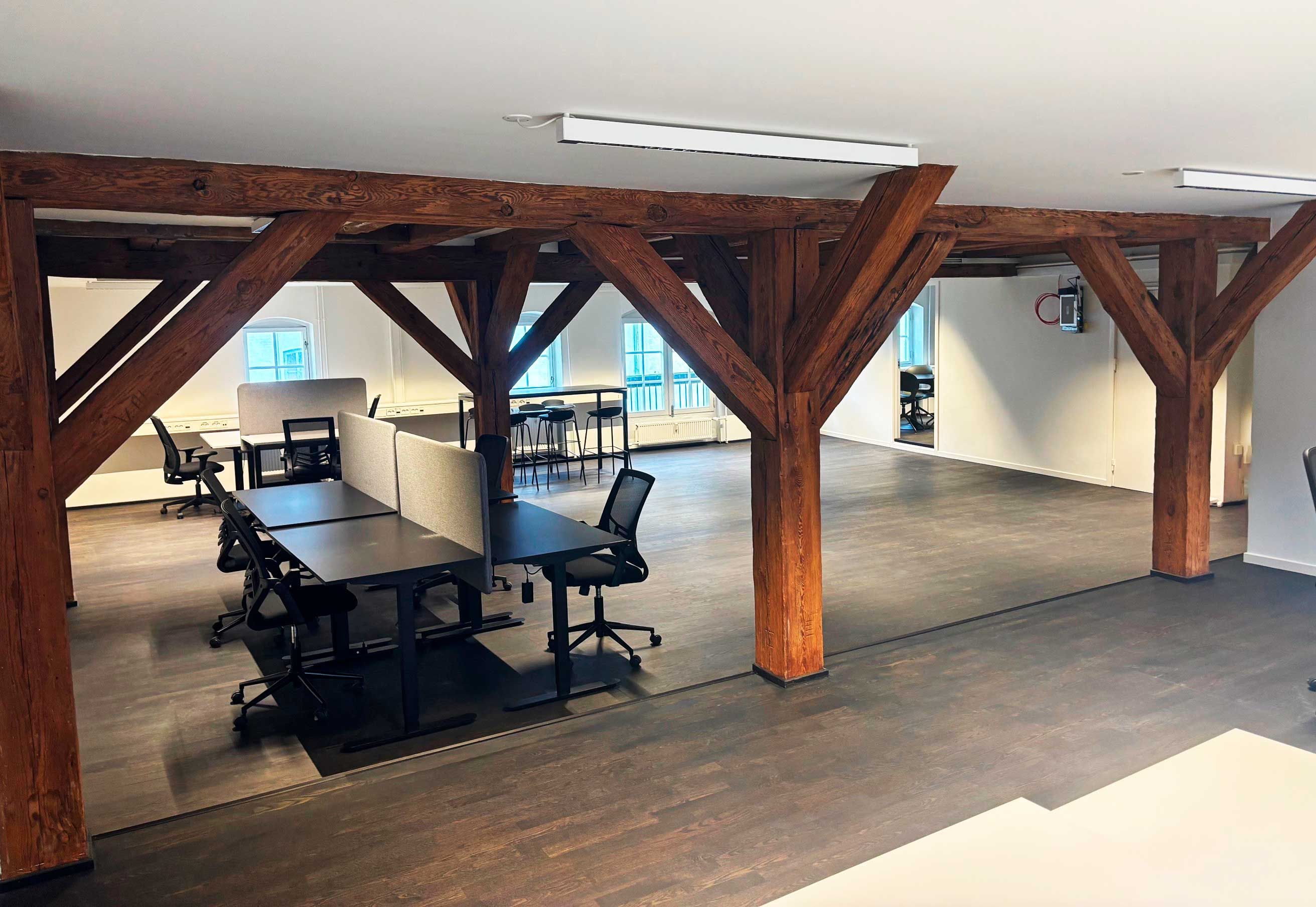 Nyrenoverede kontor med træbjælker, højloft og moderne møbler. Bord, stole og skriveborde er placeret rundt om i lokalet. Der er også flere vinduer og slukkontakter.
