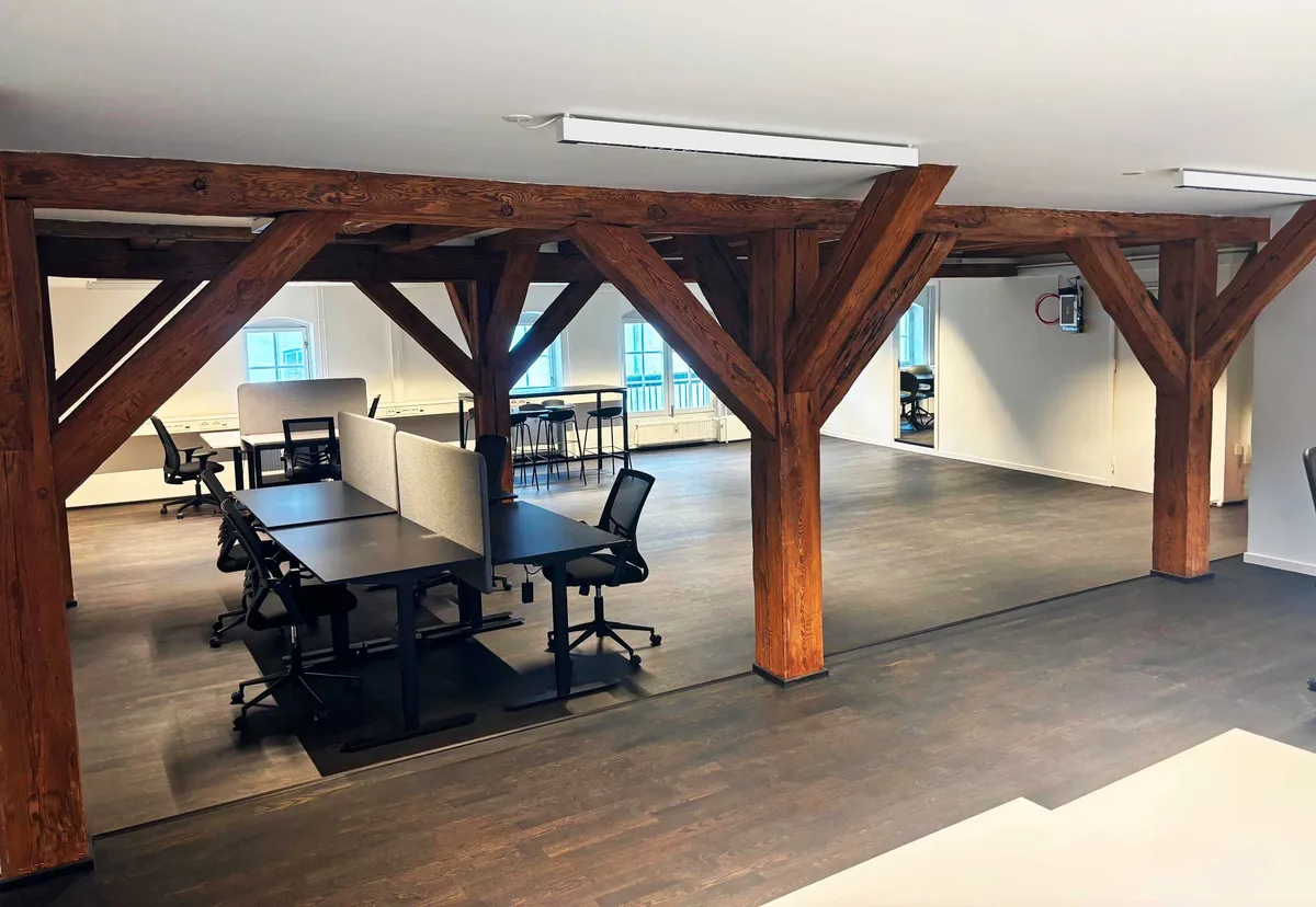 Nyrenoverede kontor med træbjælker, højloft og moderne møbler. Bord, stole og skriveborde er placeret rundt om i lokalet. Der er også flere vinduer og slukkontakter.