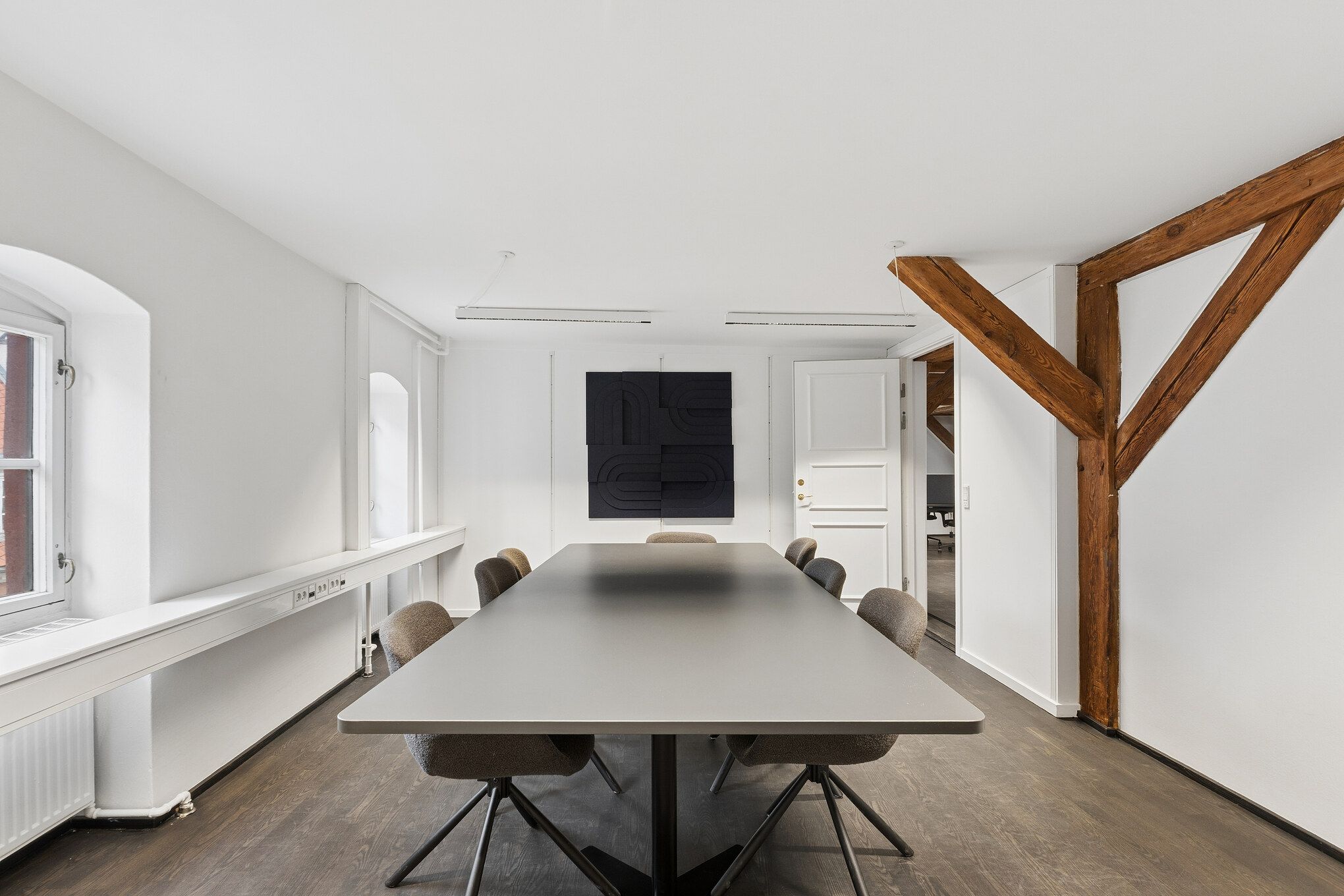 Moderne kontor med lyse vægge og lysekroner, en lang bord med stole og et sort maleri på væggen i lysegrå farve. Vinduer med lysegardiner og en åben dør