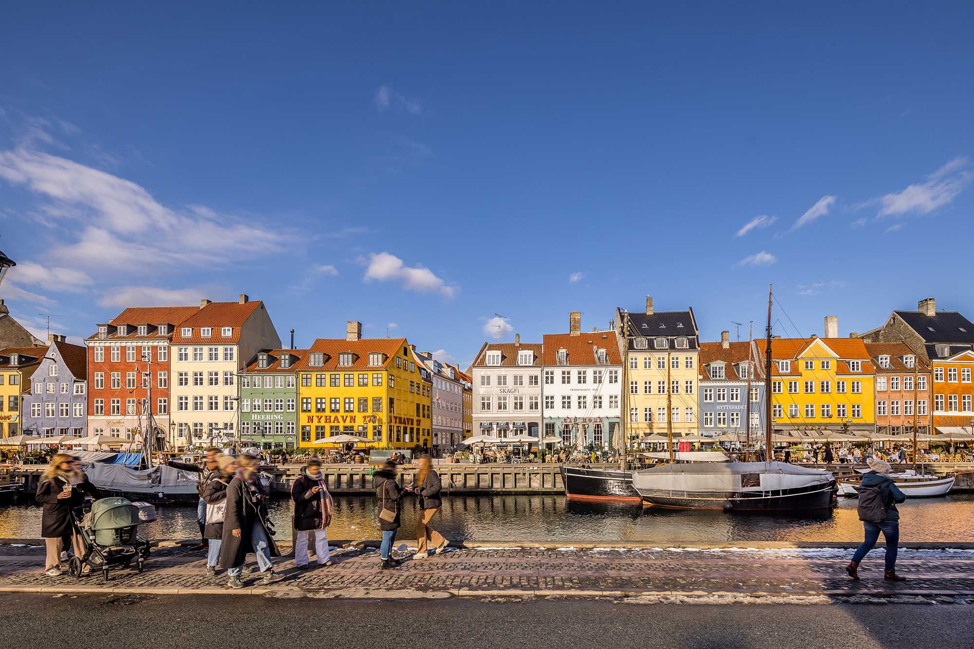 Folk går på hjørnet af Nyhavn og Ofelia plads, med farverige bygninger i baggrunden og en båd i vandet
