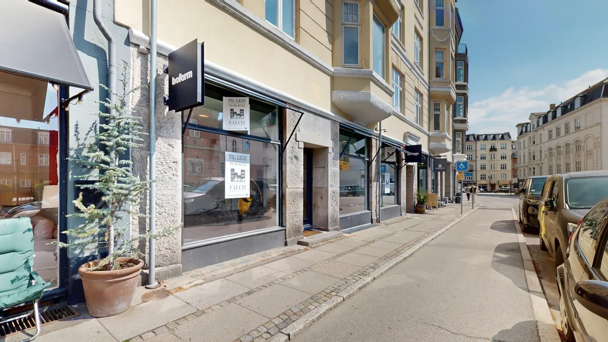 En moderne butikslås med mærkeskilt og et indvendig udsalgsområde med møbler og plante. På gaden er der parkerede biler, og der er udsigt til en anden bygning.