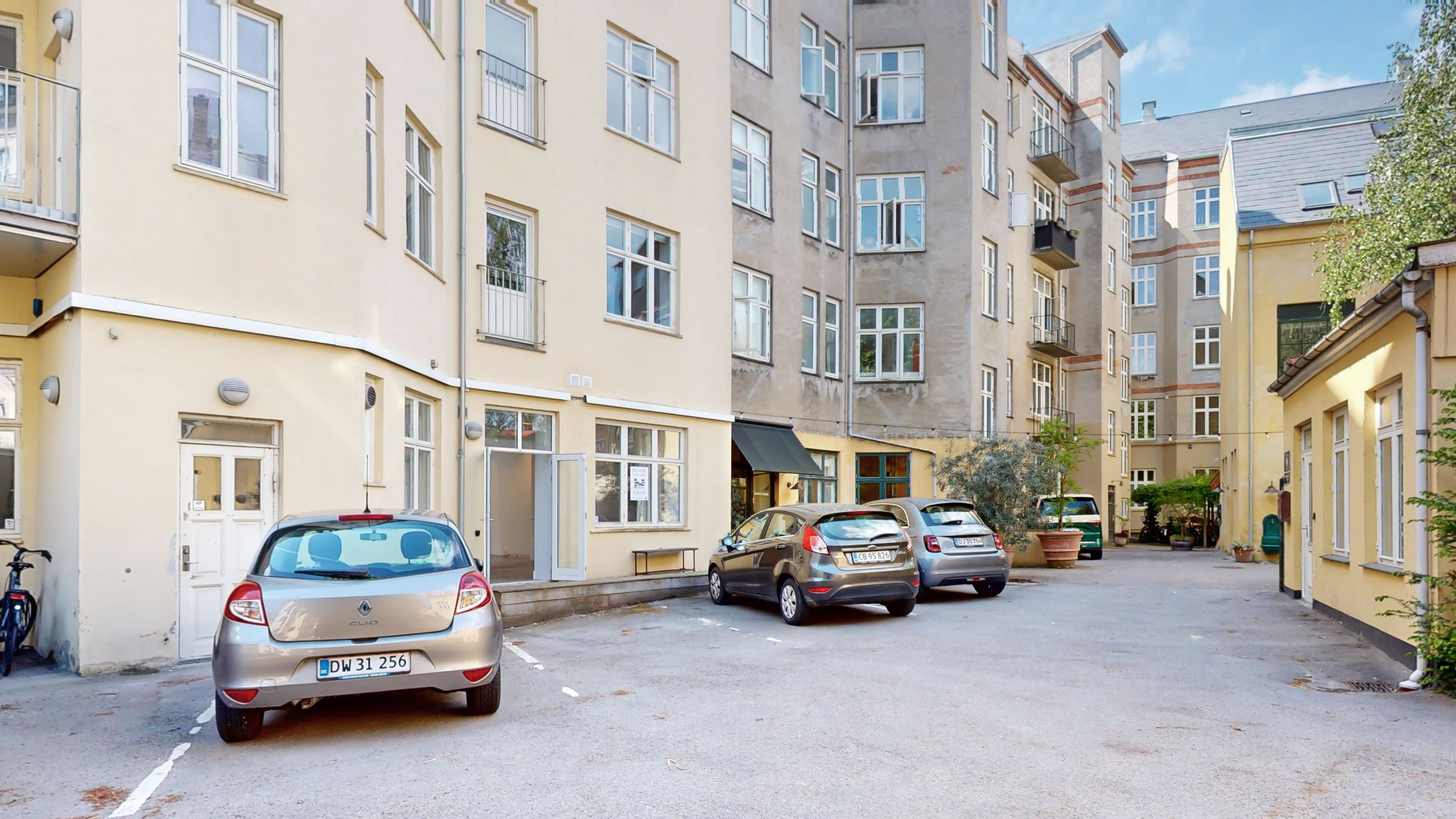 Lejlighedsbygning med fire parkerede biler og en cykel parkeret foran. Bygningen har gule vægge og flere vinduer og balkoner.