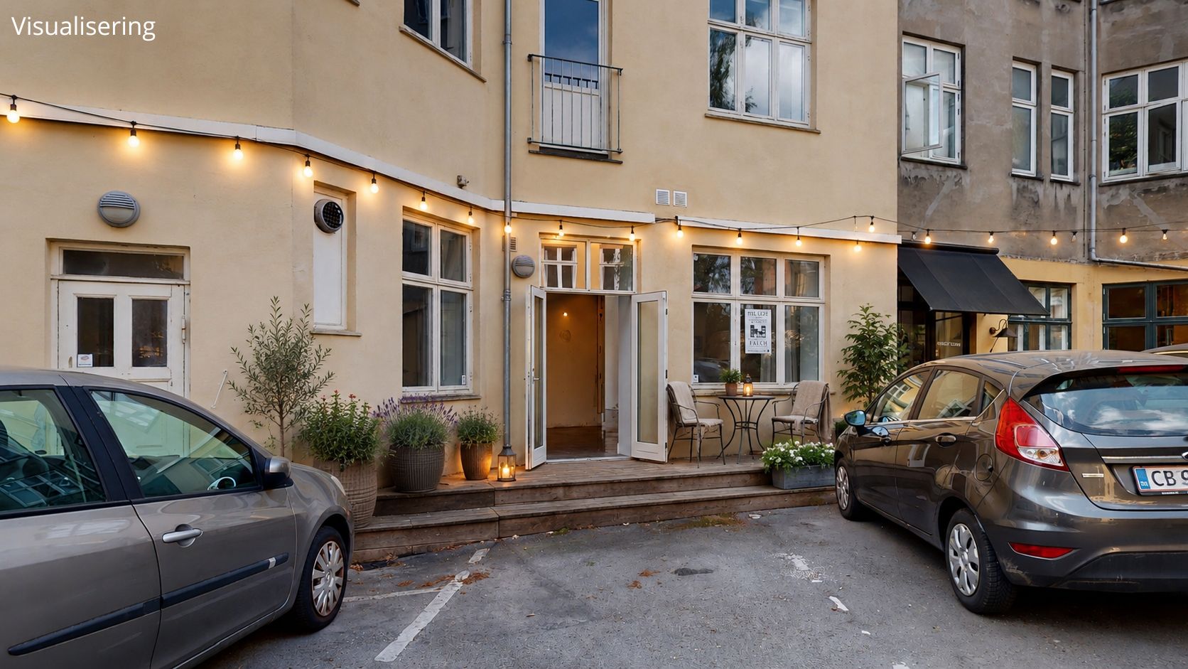 To biler parkeret foran et hus med en mørk avisstand og en indgang med lysekrans. Stiger med plantepotter og blomster samt en rundt bord med stole.