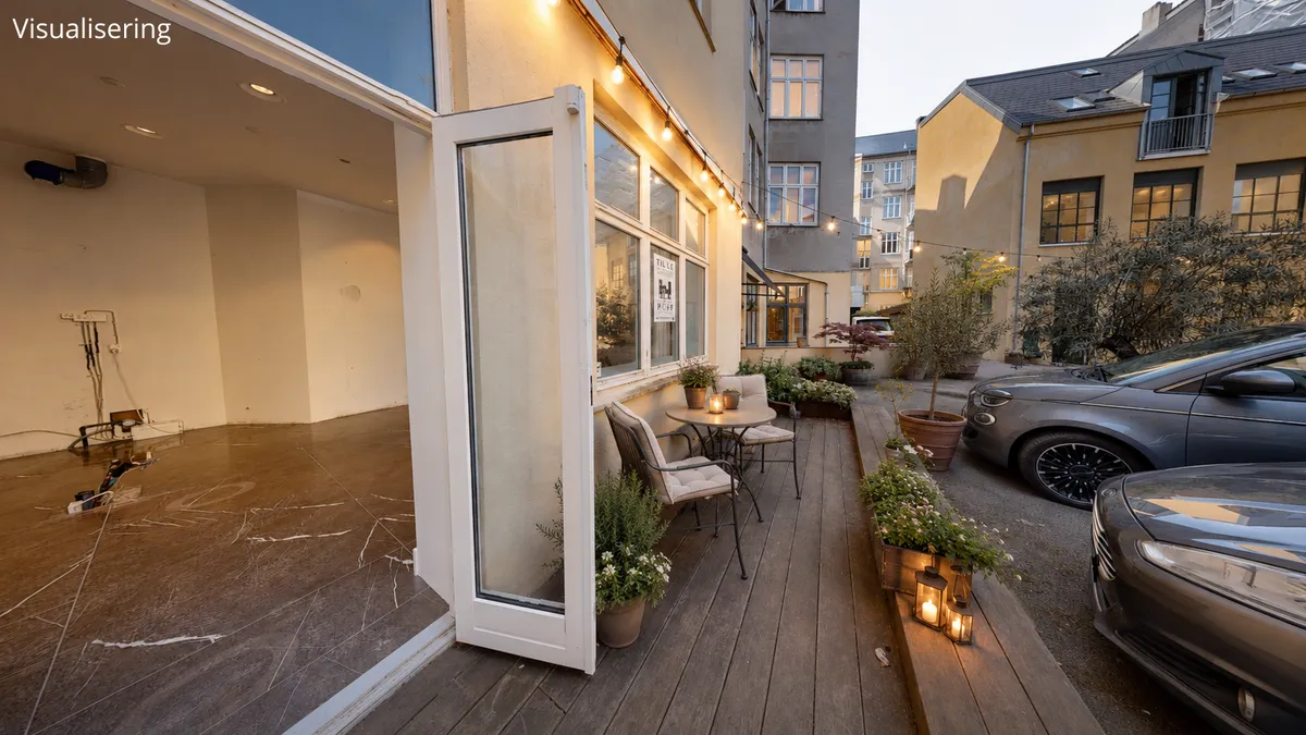 Billede af et spisestue med indgangsdør åben, udendørs terrasse med bord og stole samt planter og lysekranse. Foran er to parkerede biler og flere bygninger med vinduer