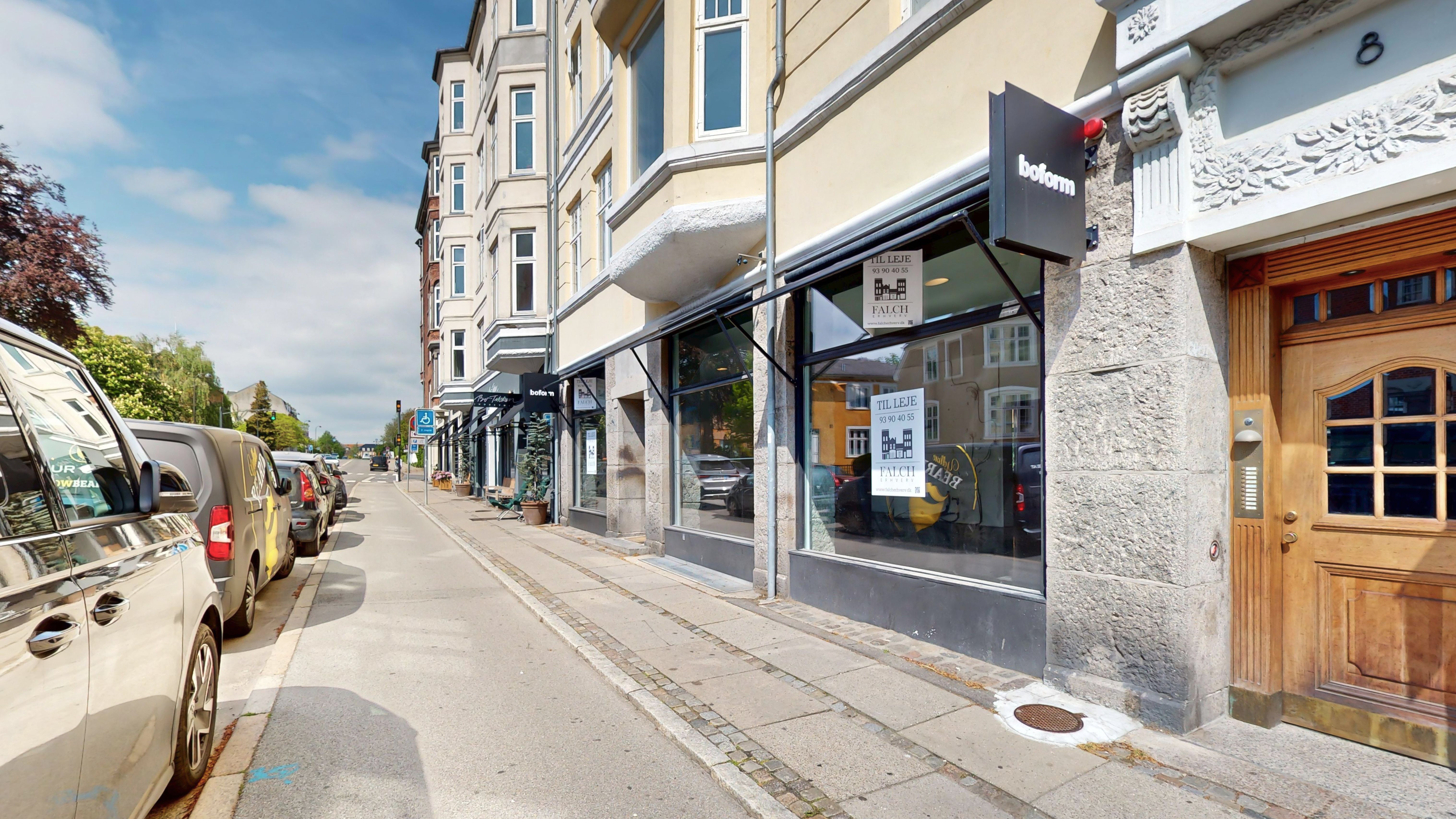 Butikslokaler til leje i et smukt gult bygningsværk med moderne facader og glasvinduer. Butikkerne vender mod en tomgangsvej med parkerede biler og udsmykkede facader