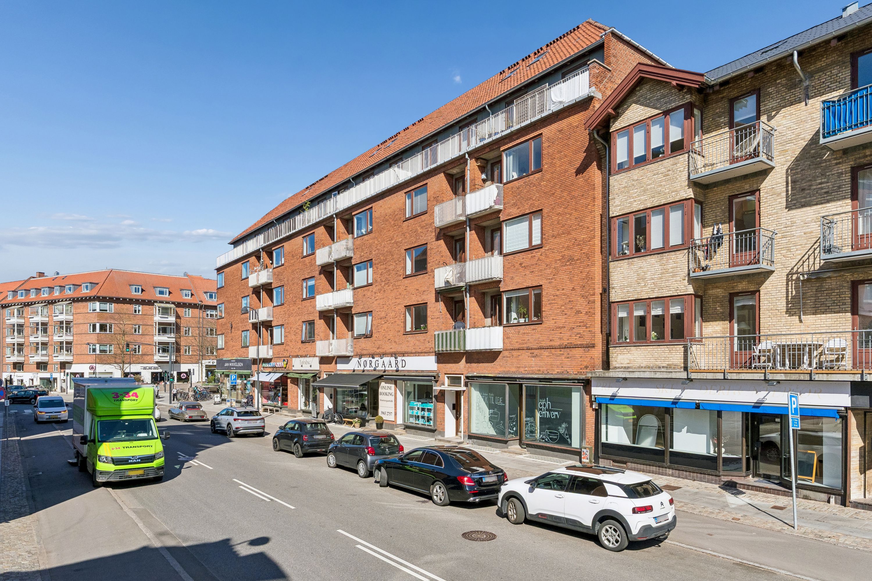 Butikslokaler med eksponeret vinduesfront på Baunegårdsvej 3, Gentofte. En stor transportvogn og biler kører på vejen. Tre parkerede biler står på højre side.
