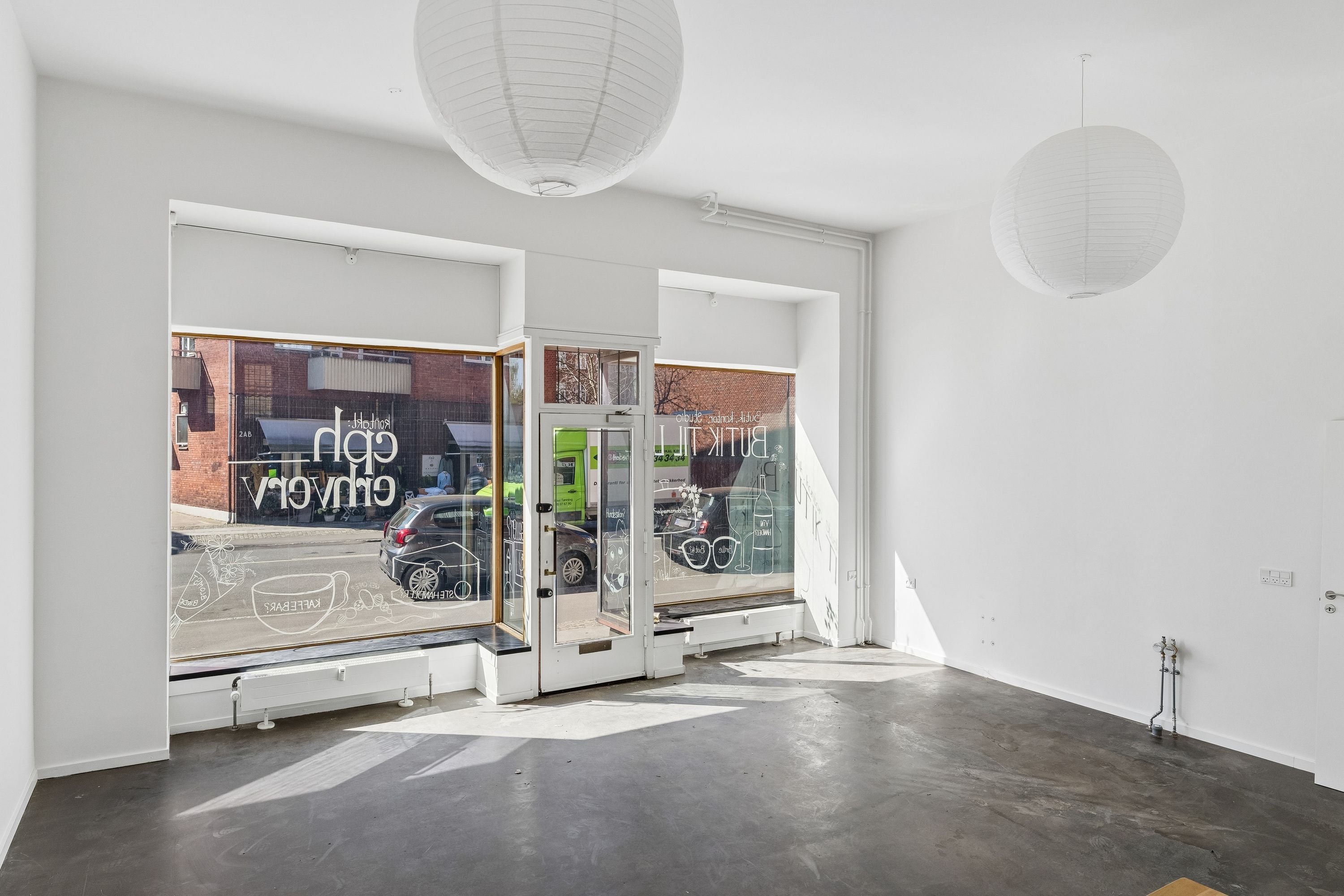Lejestue med to papirlampelys, hvide vægge og gulve samt store vinduer. Der er en glasdør med tekst og tegninger og to radiatorer i højre sidevæg.