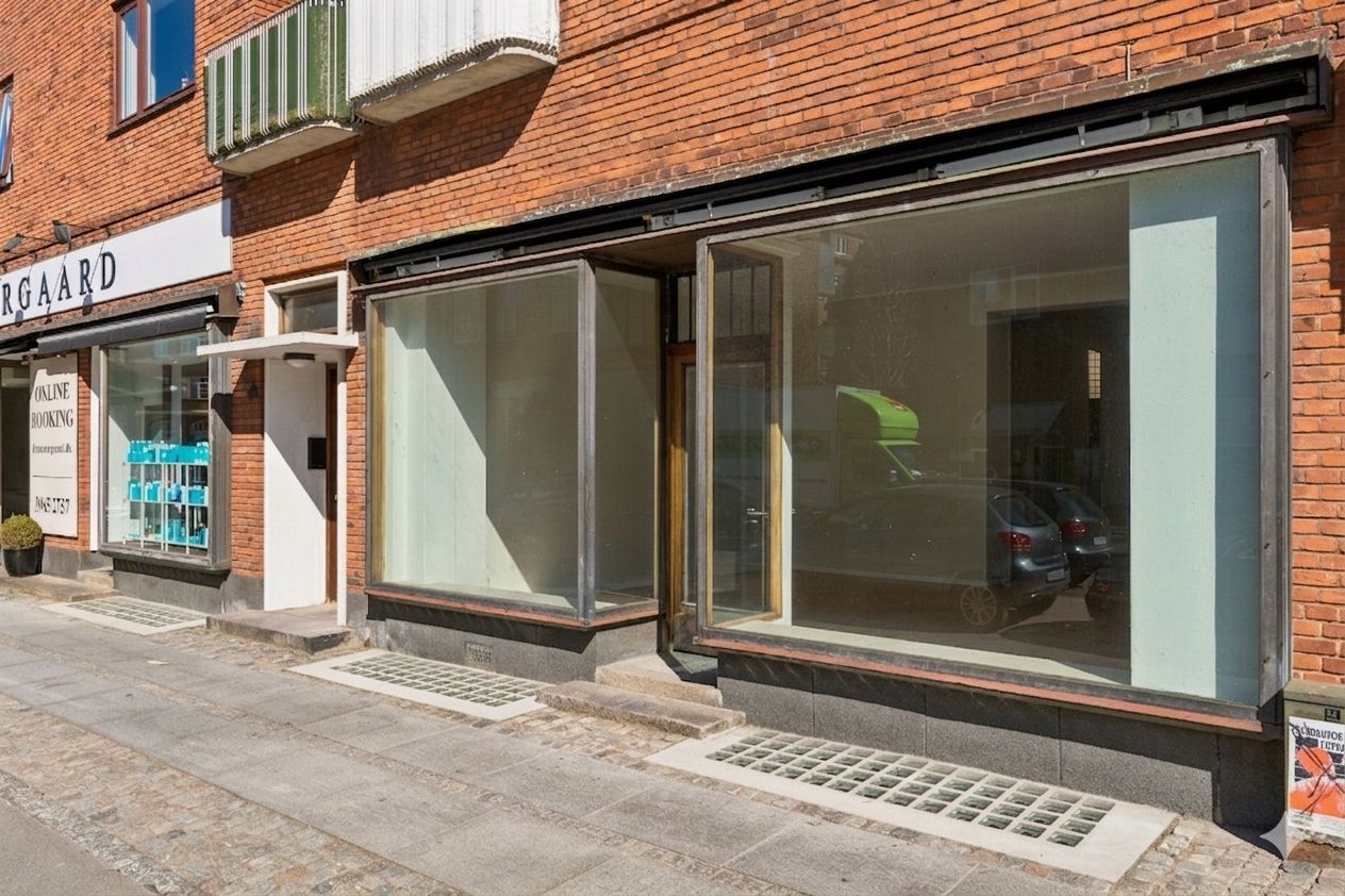 To butikker med vinduer og reklameskilte. En med en hvid facade og en med røde mursten. En plante i potte og flere biler parkeret. En hvid skrånende overgang til en fortovskant.
