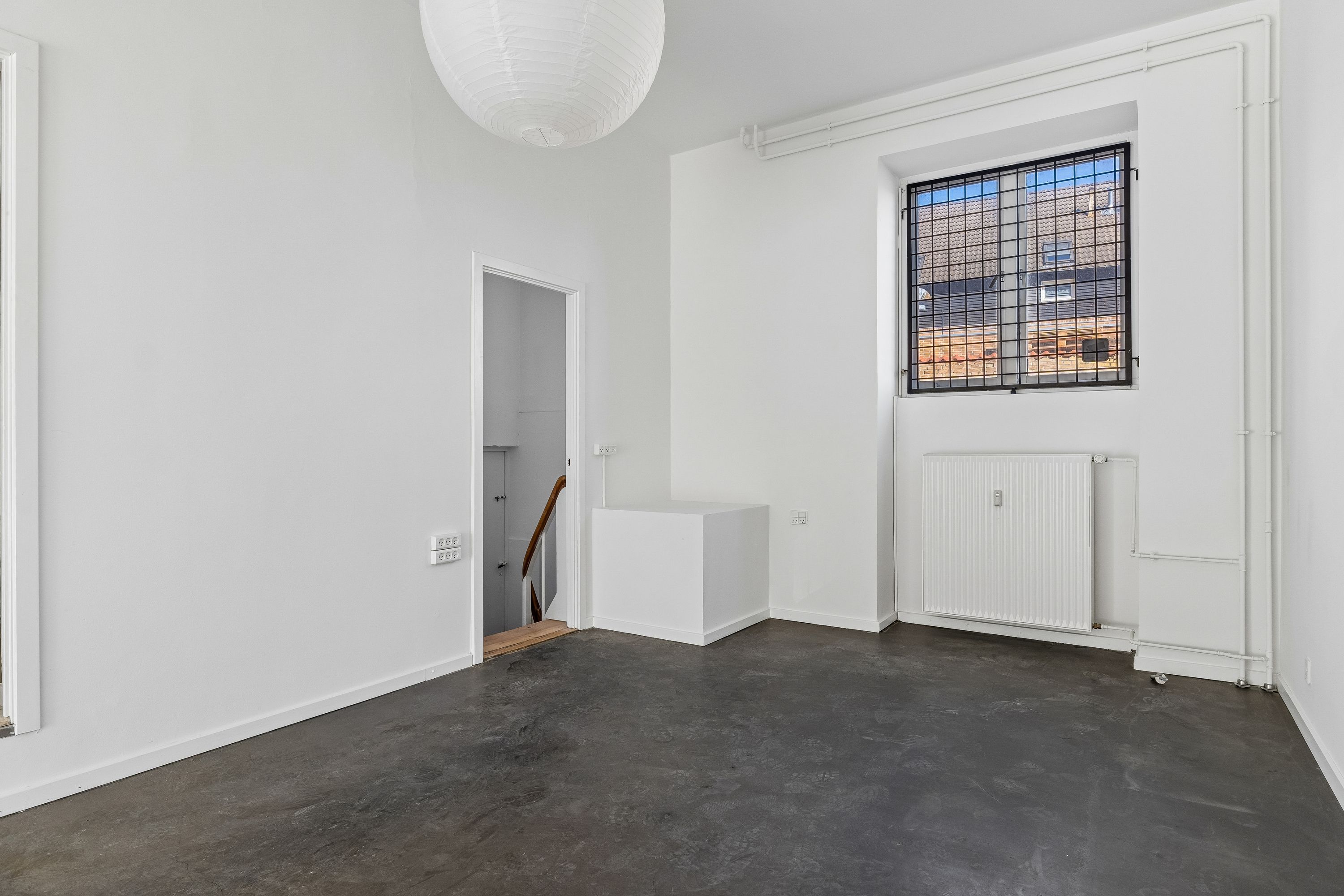 En møbleret lejlighed med hvidt loft og vægge, hvor der er en radiator og en hængende lampeklokke. Der er et vindue med skur og et åbent område med trætrappe.