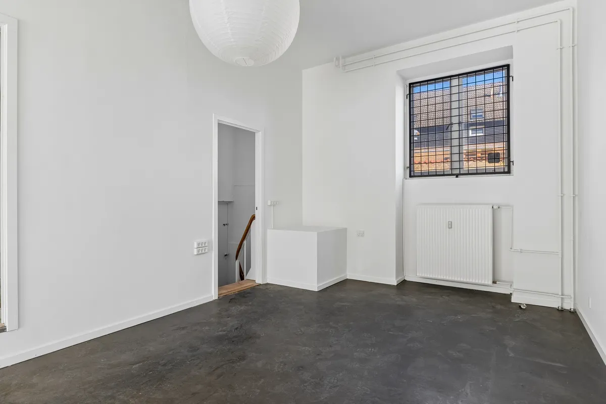 En møbleret lejlighed med hvidt loft og vægge, hvor der er en radiator og en hængende lampeklokke. Der er et vindue med skur og et åbent område med trætrappe.