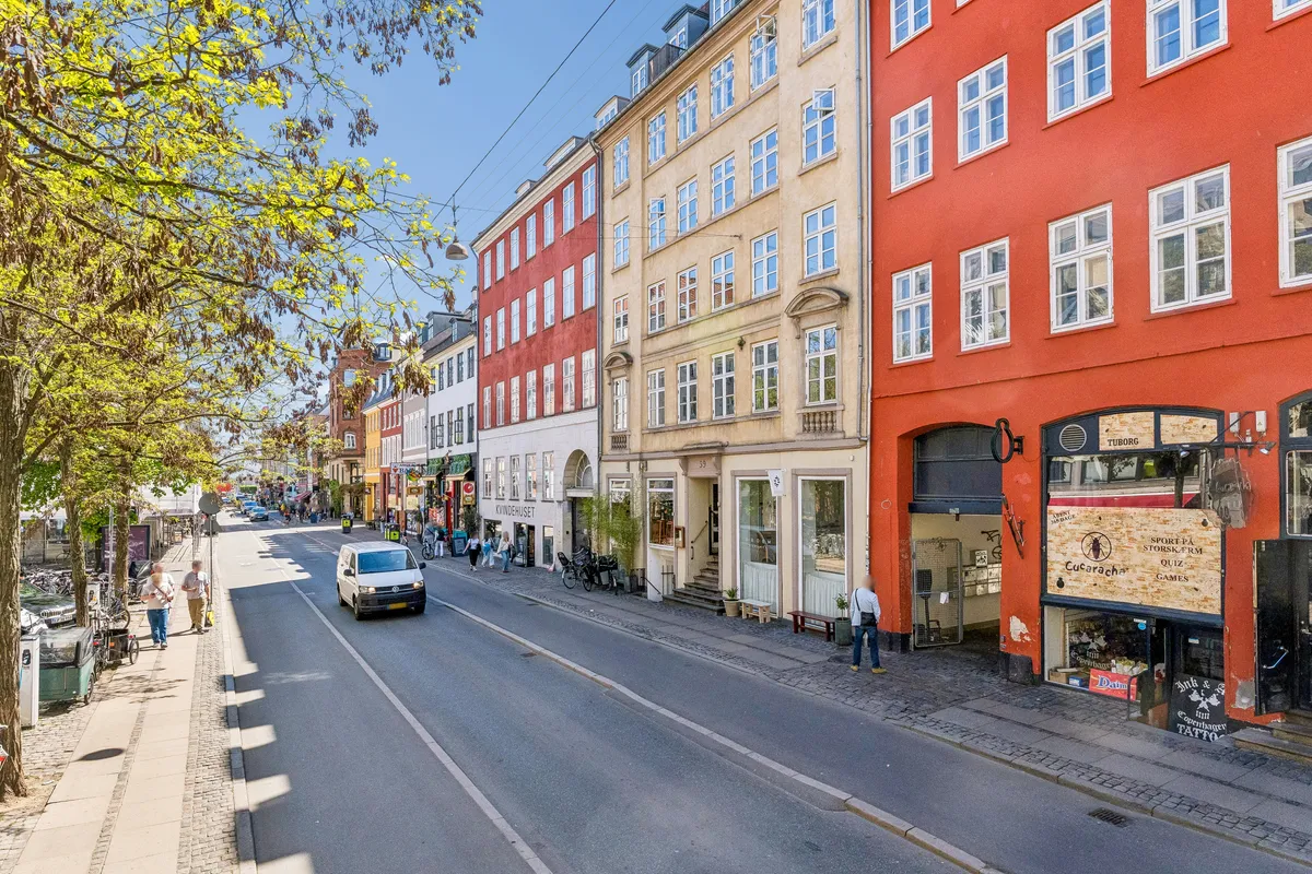 Butikstilbud på Gothersgade i København. Vandret udsigt over gågade med bil og fodgængere. Dekorative røde bygninger med vinduer og grønne træer.