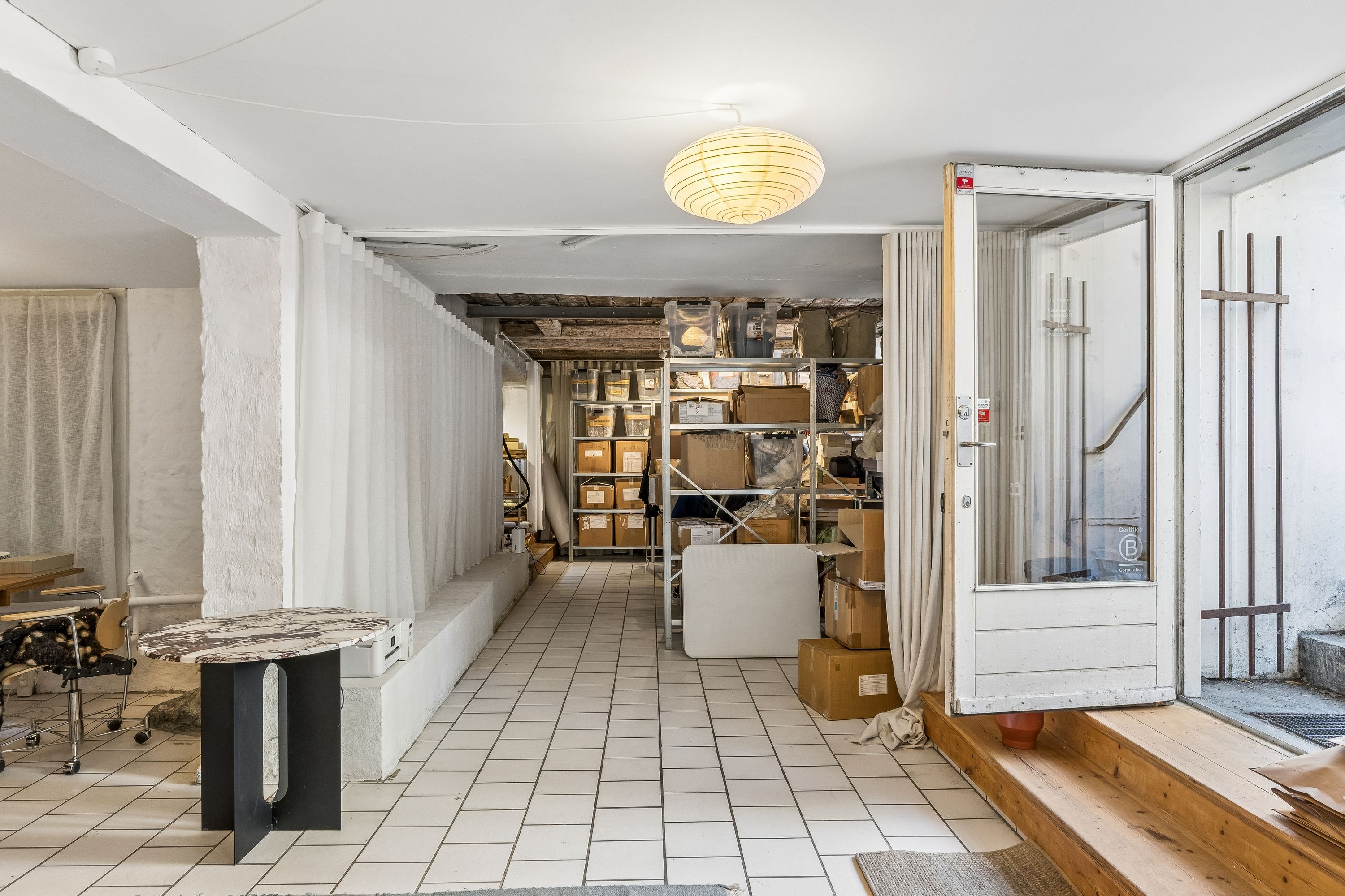 Butik med hvid interiør og møbler, stående på blankgulve og forskudte af høje lagerhylder med emballage. En åben dør og vinduesruder med hvide gardiner