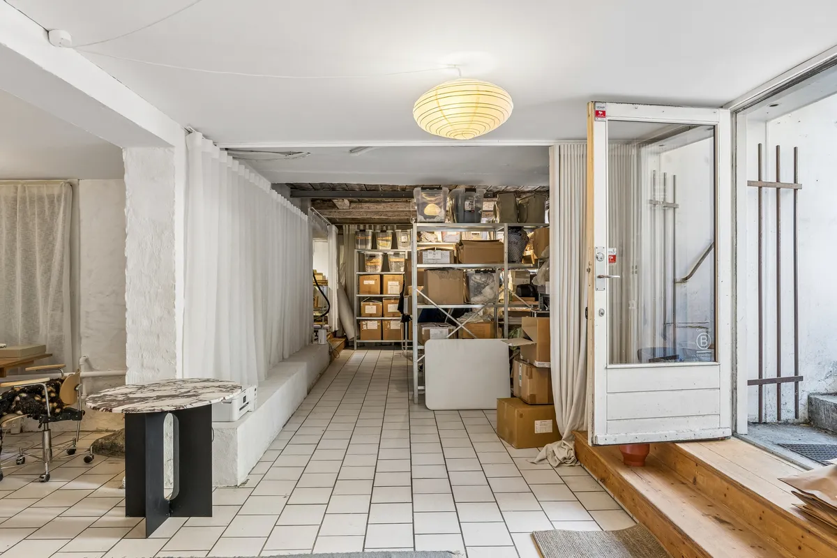 Butik med hvid interiør og møbler, stående på blankgulve og forskudte af høje lagerhylder med emballage. En åben dør og vinduesruder med hvide gardiner