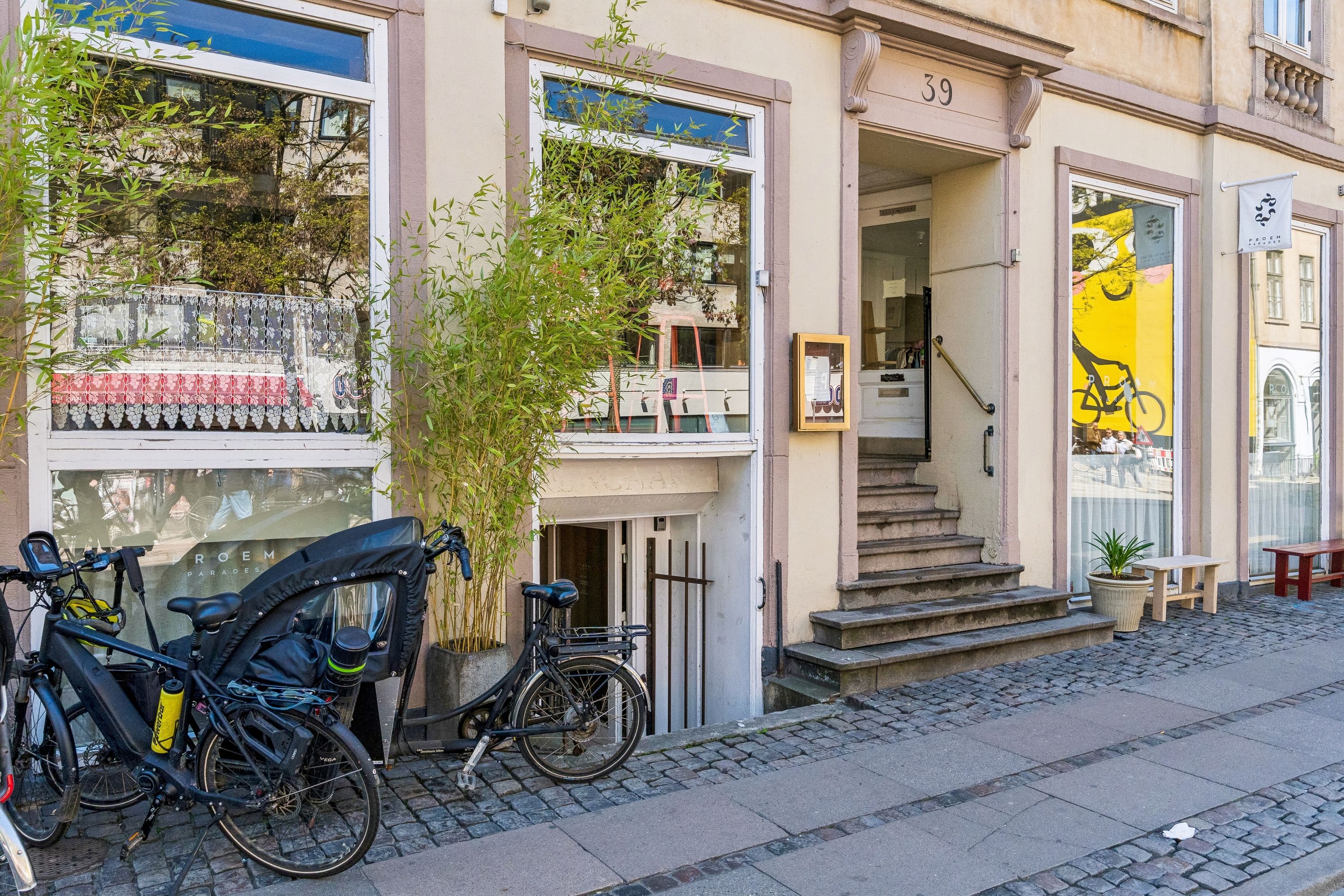 Lejlighed til leje på Gothersgade 39 med grønne planter, cykler og en gul tekst på vinduet. Butik med opgang og trapper. Adgangsforhold er let, og området er attraktivt.