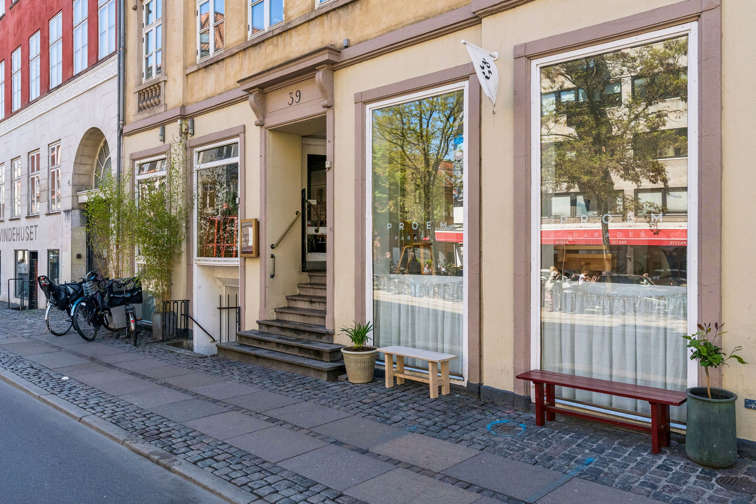 Butiksfront med nummer 39 på Gothersgade. Vindue med refleksioner af gade, træer og folk. Træbænk og plantepoter med grønne planter. Stiger med bjælke. To cykler parkeret.