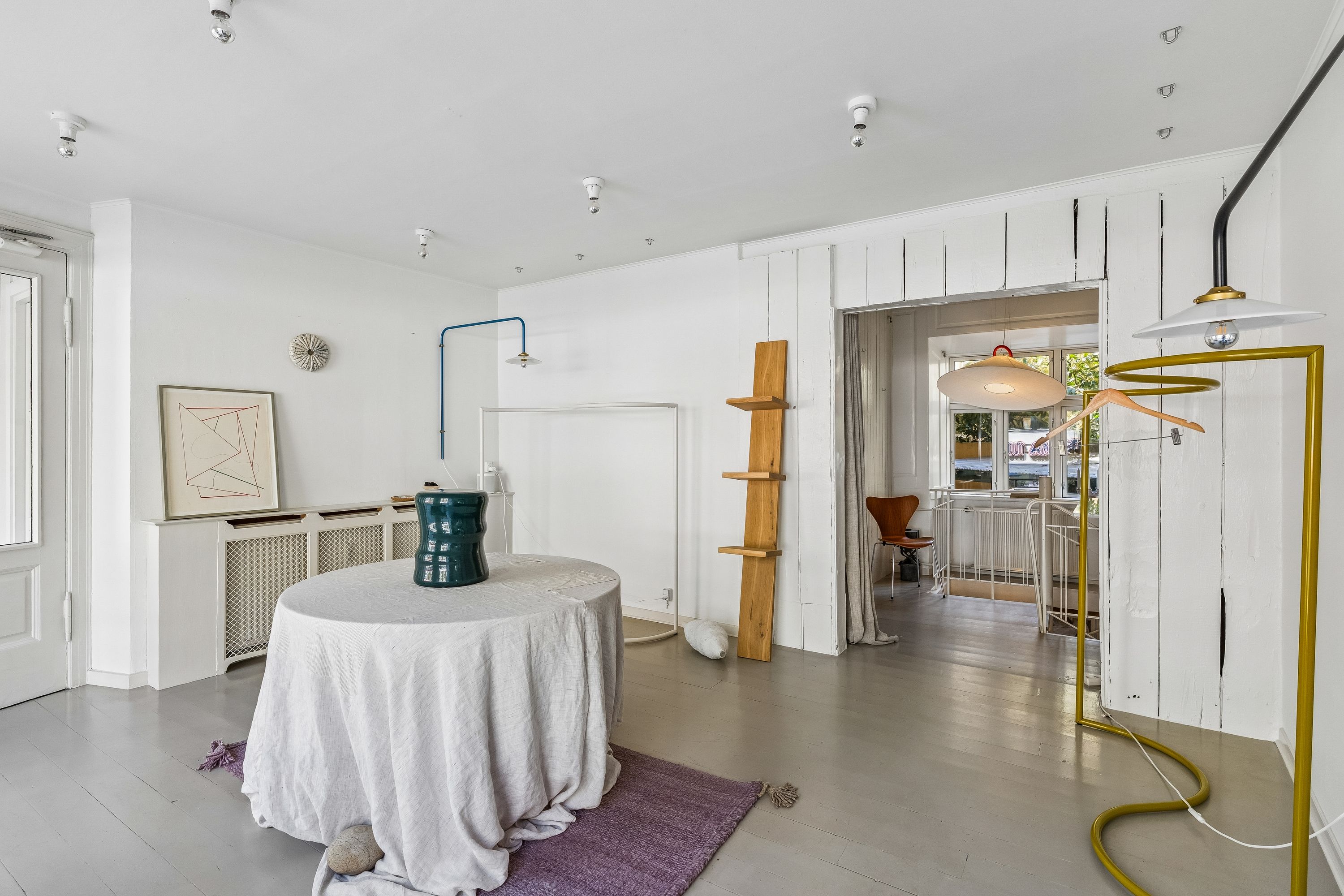 Lejlighed med minimalistisk indretning og hvidt interiør, stol ved vinduet, hængende lamper og et hvidt bord med en grøn vase og en klud der dækker det