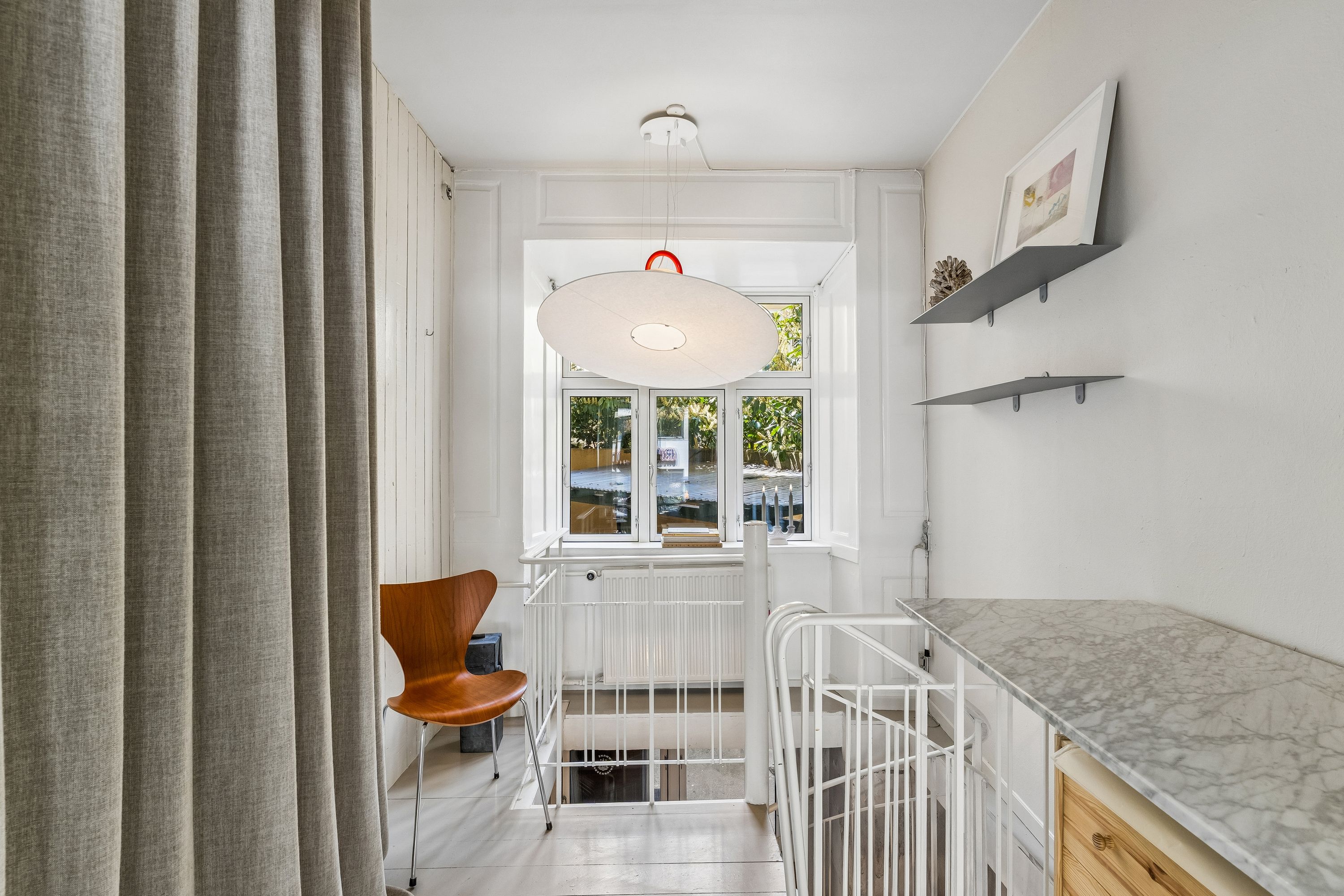 Indvendigt moderne møbler med lyse gulve og lysegrå vægge, hvidt loft med lysekron, høj vinduesåbning med udsigt til gaden og møbler og hylder med kunstværker.