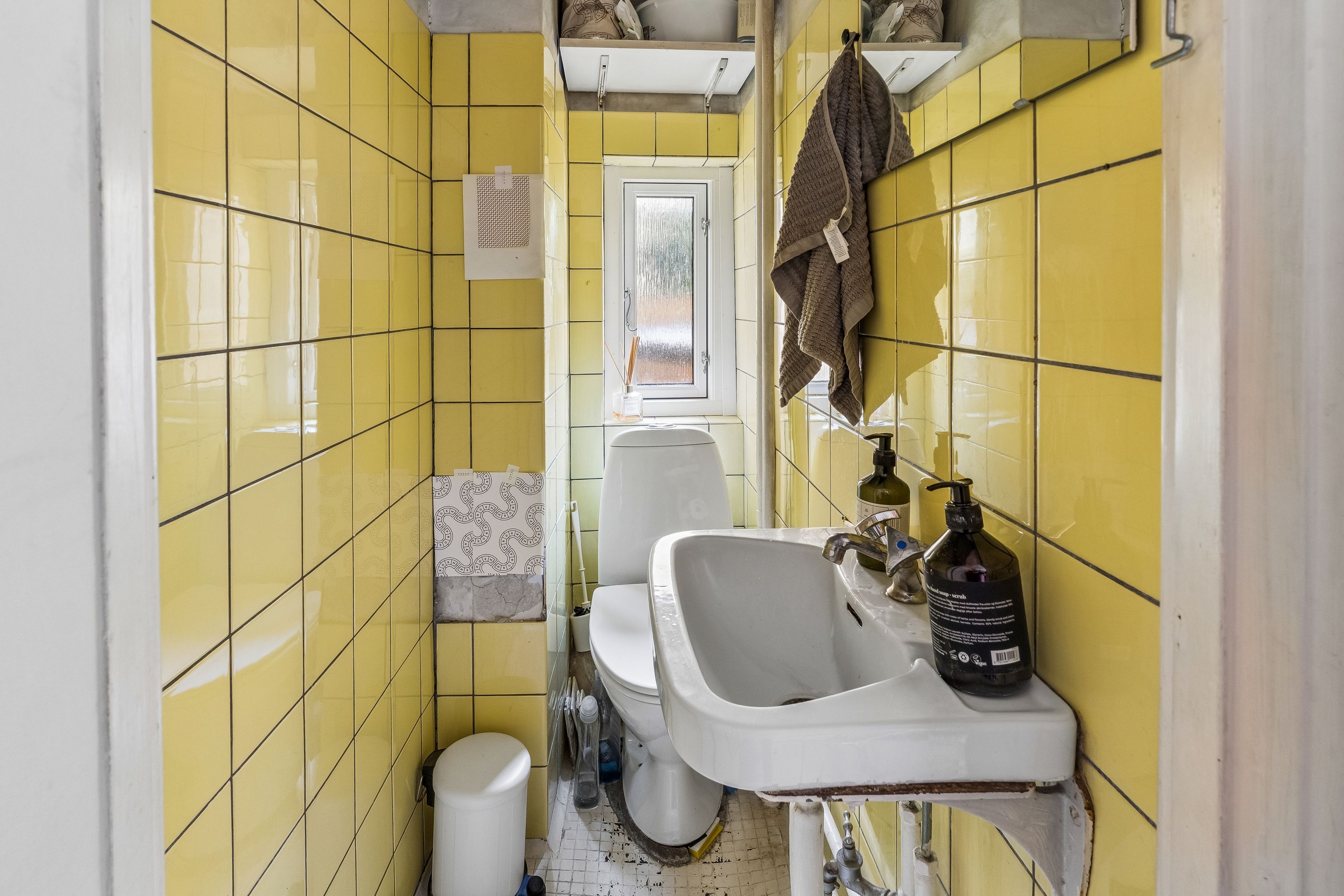 Et lille badeværelse med gule fliser og et toilet med hvid skål og briller. En håndklæde hænger på væggen. En hvid vask med en flaske og en sæbeflaske.