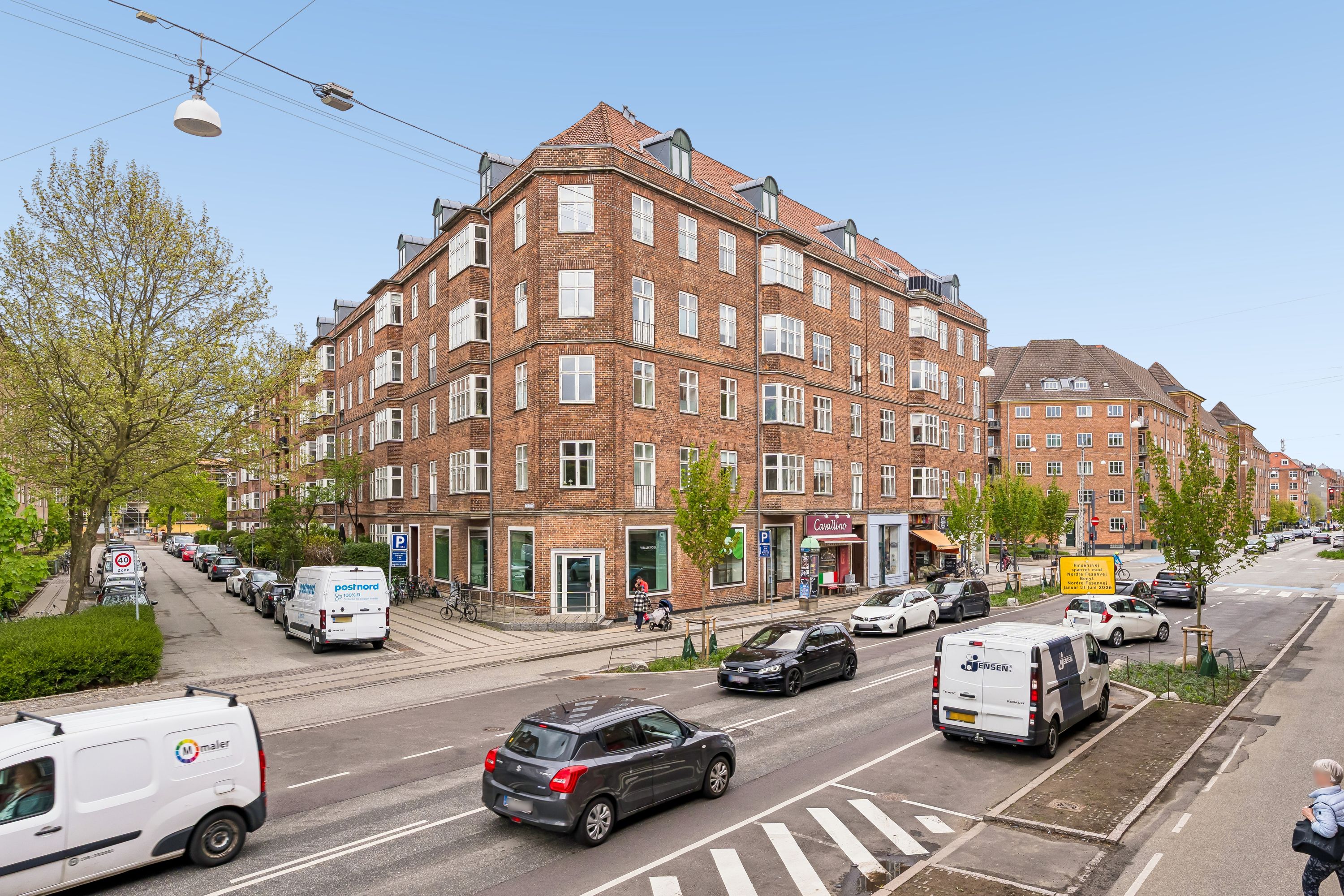 En hjørnebygning med mange lejligheder og butikker, hvor biler og folk er på gaden og en kvinde går på fortovet