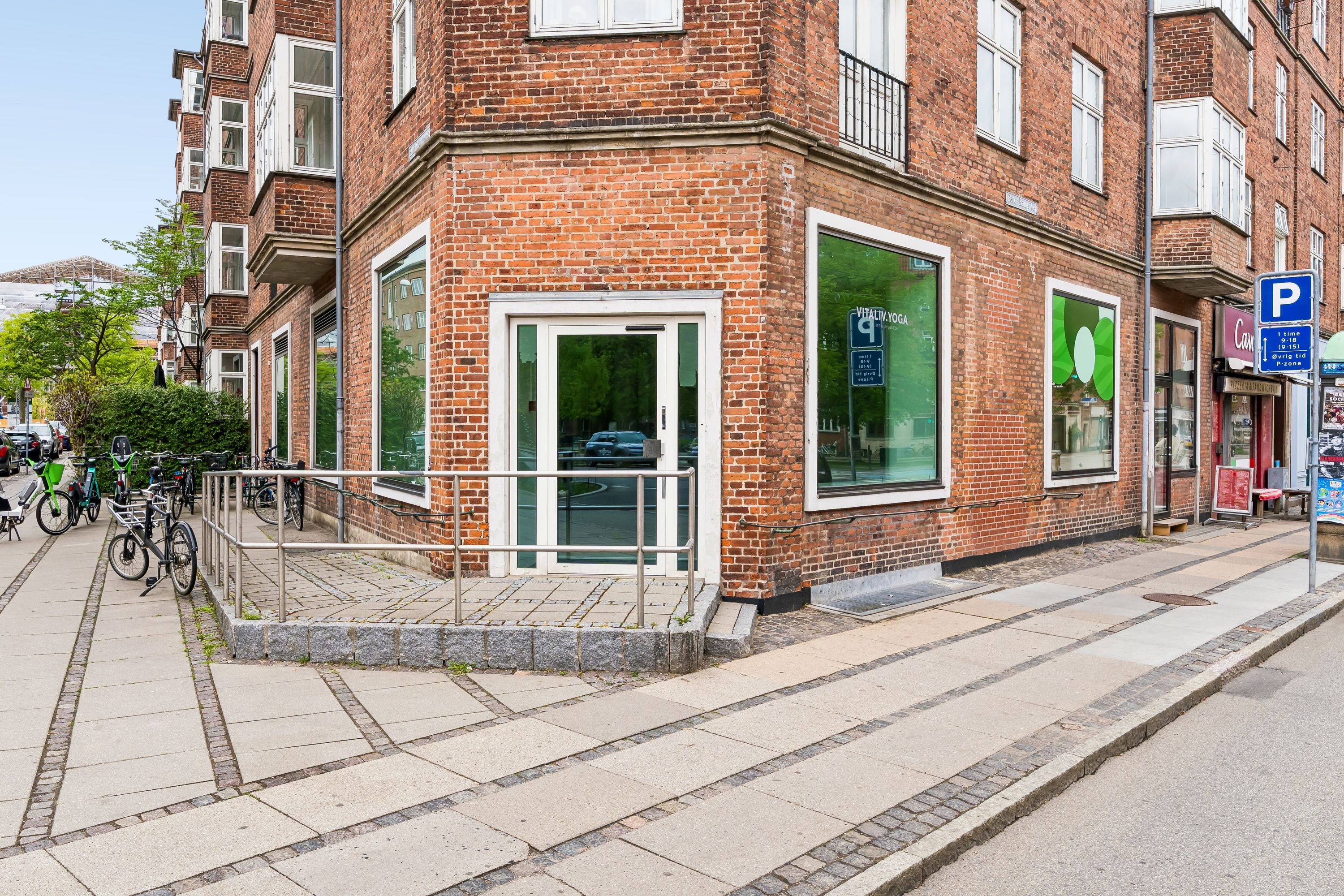 Hjørnelejemål på Peter Bangs Vej 60 i Frederiksberg. Moderne butikslokaler med glasdør og vinduer. Adgangsrammer og rulkærme på fortovet. Adgangsvej med parkerede cykler. Parkerede biler og grønne træer på gadeniveau.