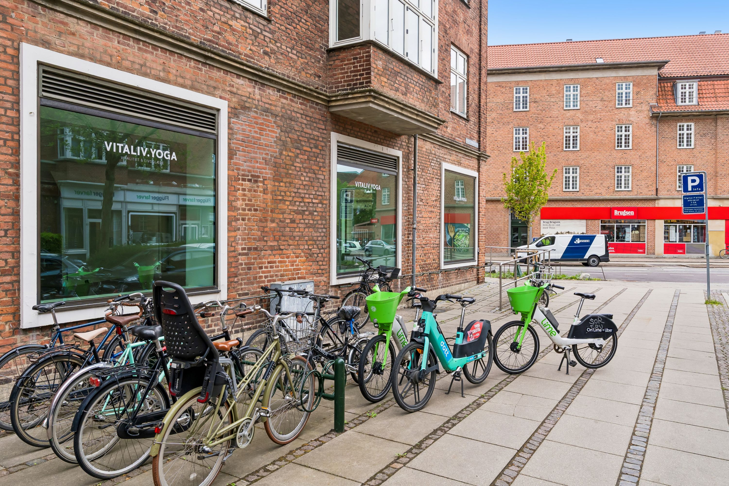 Hjørnelejemål på Peter Bangs Vej med lejecykler parkeret foran. Yogacenteret VitalIV Yoga ligger på hjørnet. Der er et parkeringsskilt og Brugden butik i baggrunden