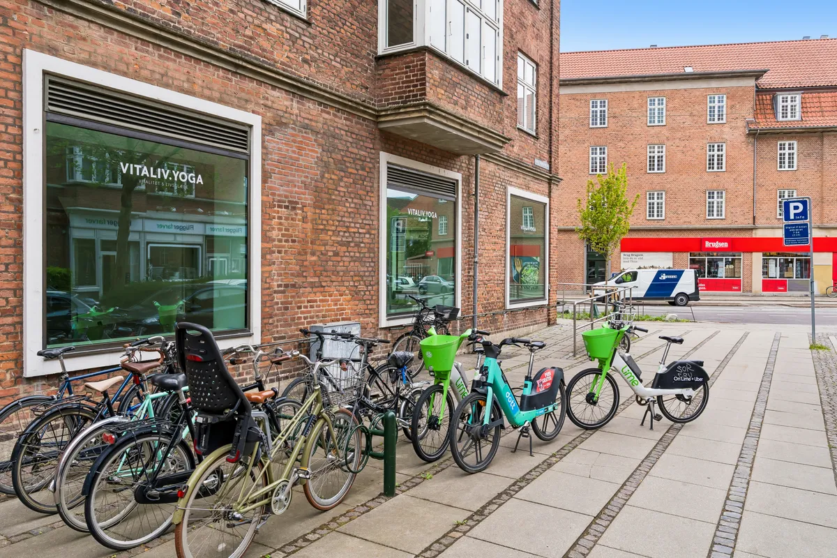 Hjørnelejemål på Peter Bangs Vej med lejecykler parkeret foran. Yogacenteret VitalIV Yoga ligger på hjørnet. Der er et parkeringsskilt og Brugden butik i baggrunden