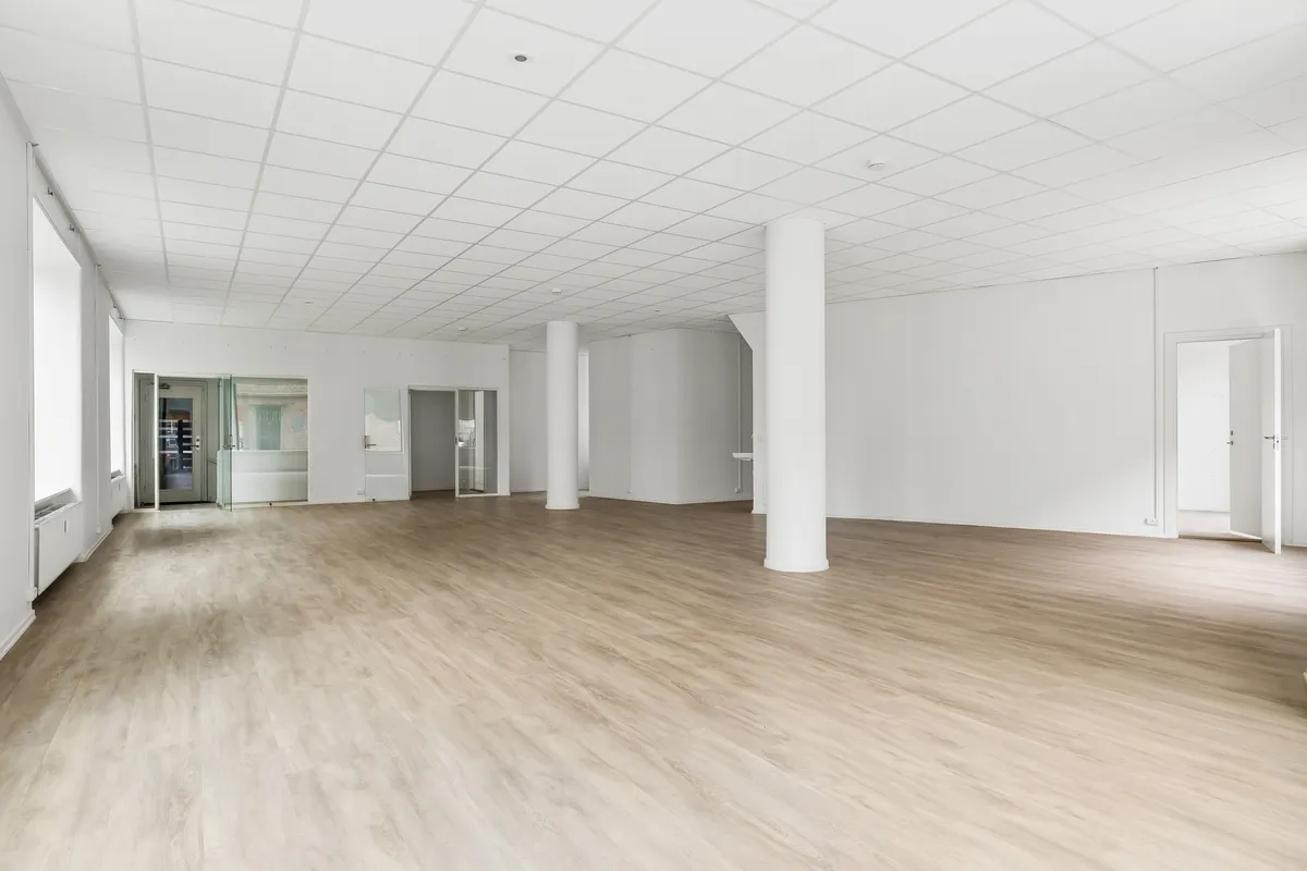 Alt: Foto af en lyse, hvidrum med trægulve og store vinduer. Der er to hvide søjler og to åbne døre til mindre lokaler.