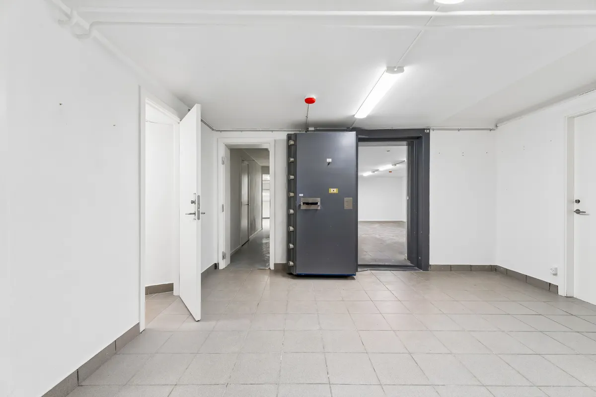 Et stort, tomt lagerrum med hvide vægge og gulv og en stor dør, der er lukket og en anden dør, der er åben. To små søjler og en dør i baggrunden