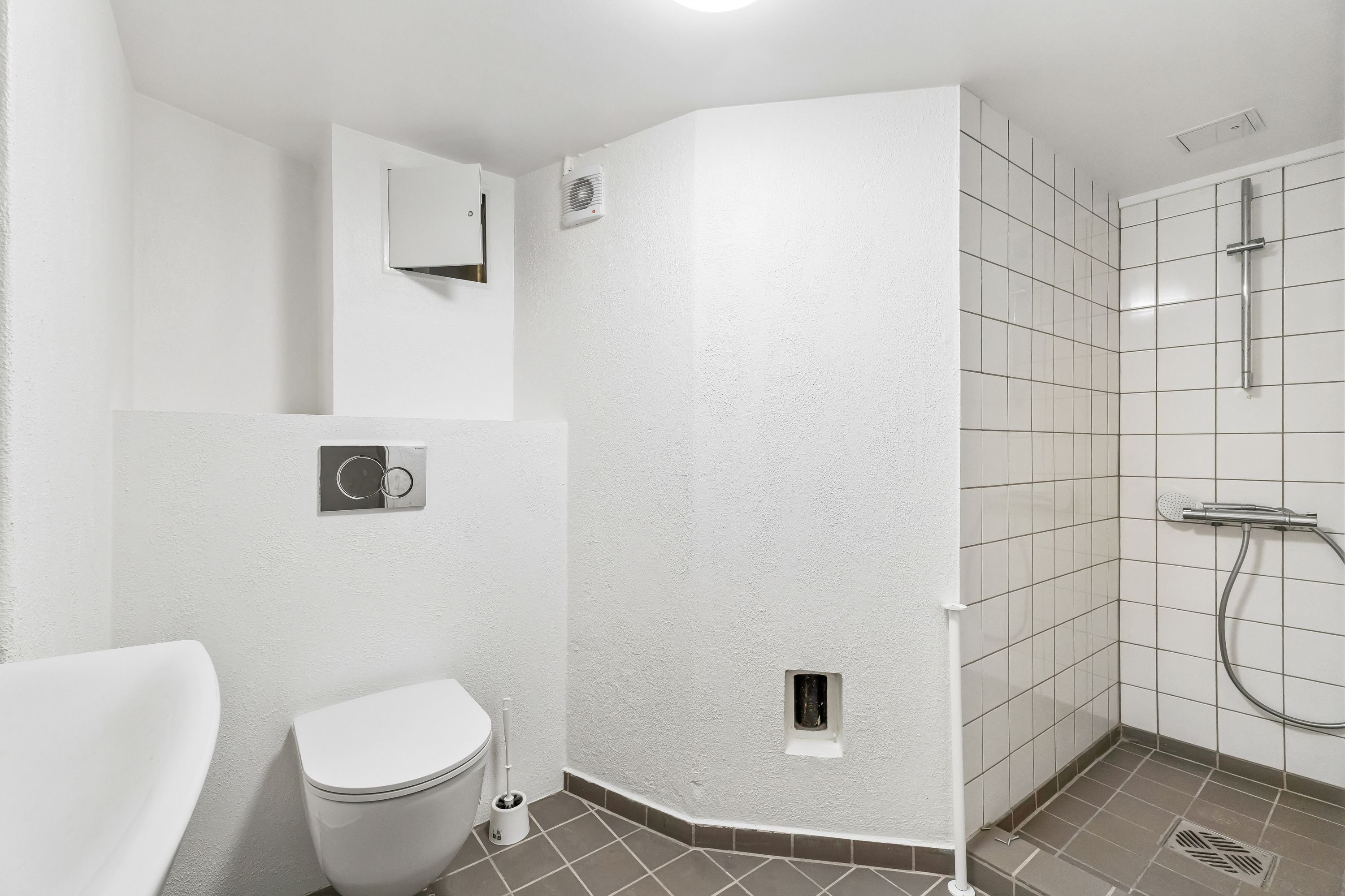 En lille, ren og moderne badeværelse med hvidt toilet, hvidt badekar og badeværelseskabinet med toiletpapirholder. Der er en hvid brusekabine og en hvid brusearmatur med slange og hånddusk.