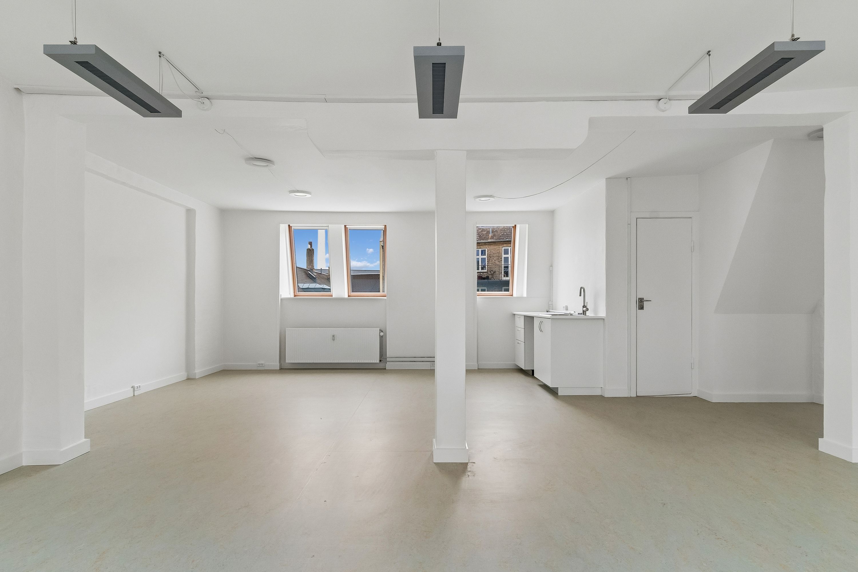 Kontor med hvidt interiør, højt til loftet, lyseruder og moderne tekniske installationer. To store vinduer mod Smallegade, hvor der ses en bygning og træer udenfor.