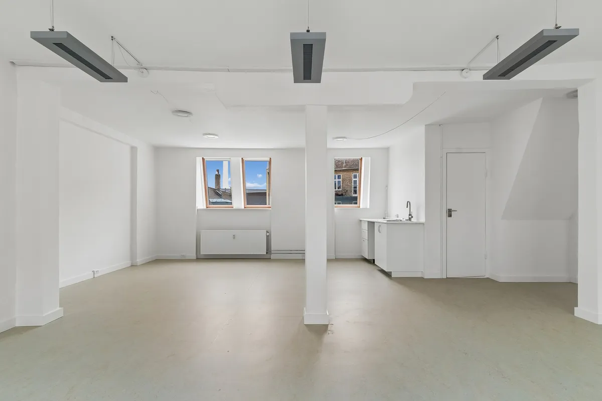 Kontor med hvidt interiør, højt til loftet, lyseruder og moderne tekniske installationer. To store vinduer mod Smallegade, hvor der ses en bygning og træer udenfor.