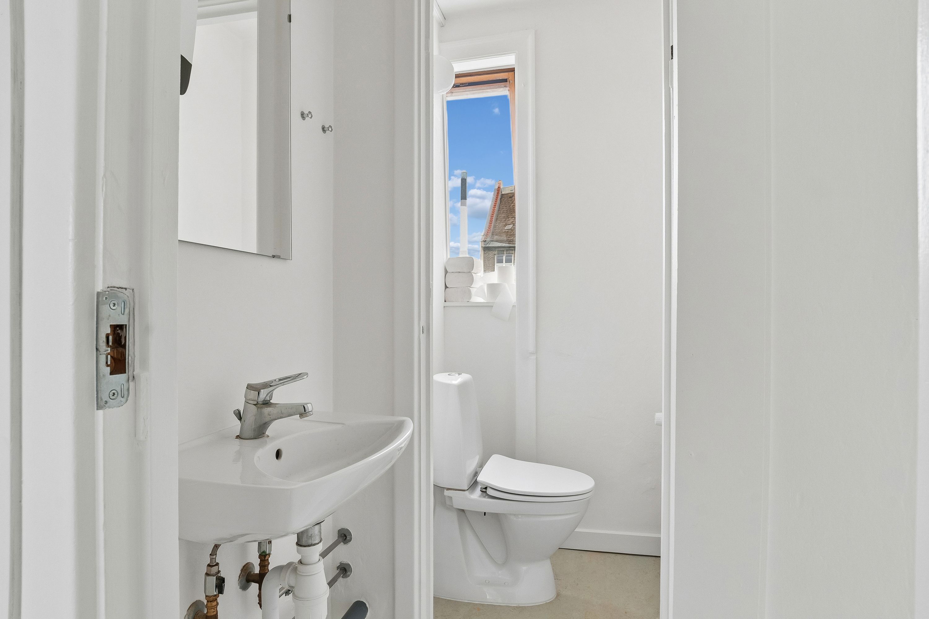 Rent badeværelse med hvidt toilet, hvidt vasketøjsblik og spejl samt lysevindu med udsigt til gavl.