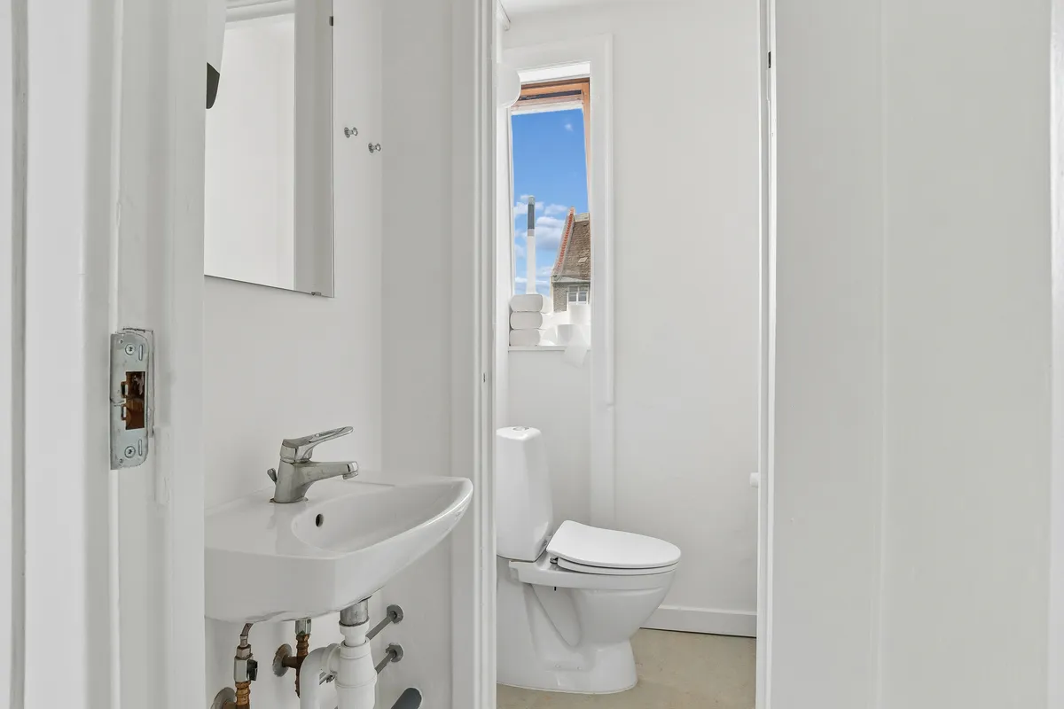 Rent badeværelse med hvidt toilet, hvidt vasketøjsblik og spejl samt lysevindu med udsigt til gavl.