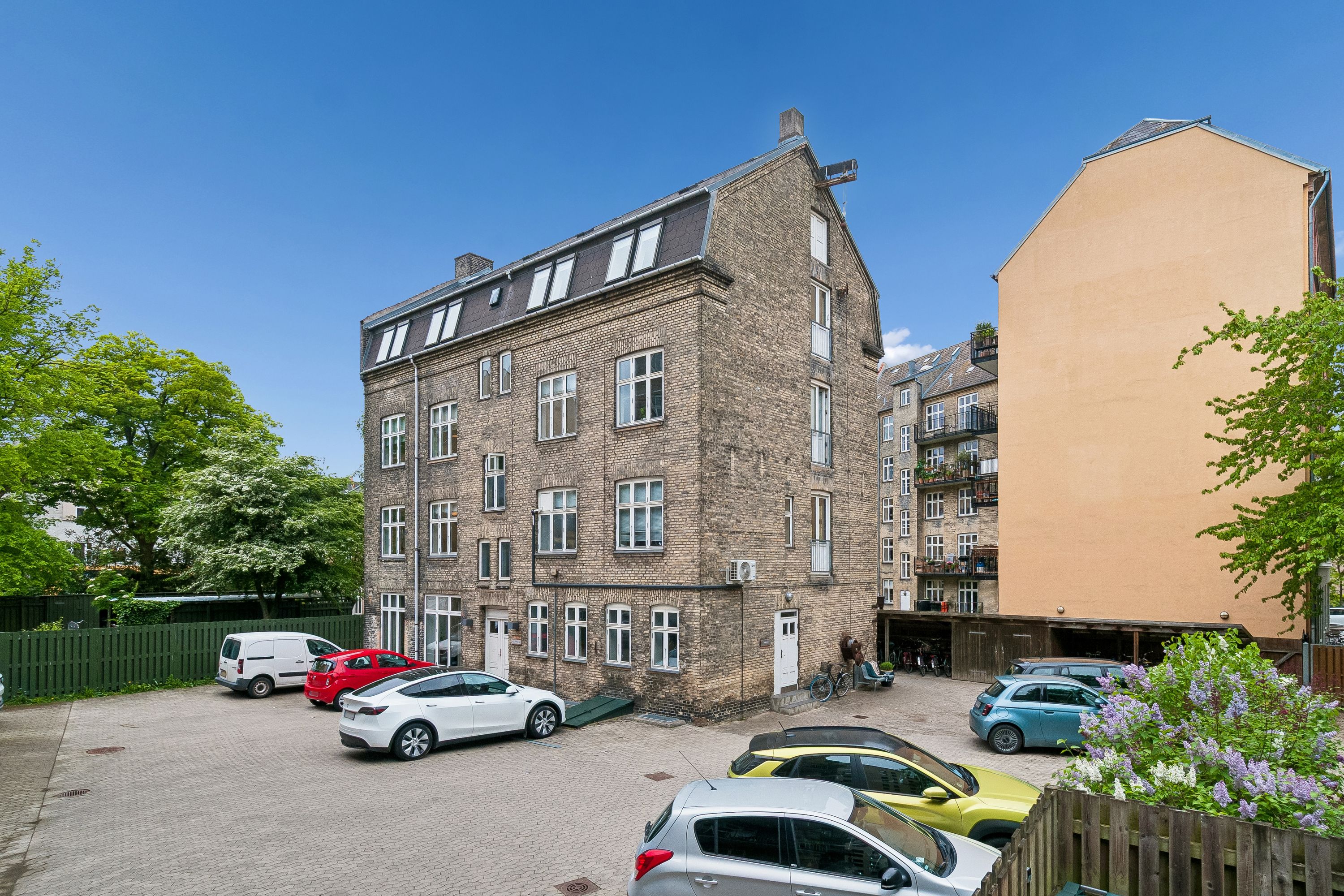 Flere biler parkeret i gården med lyse farver og lyserøde buske. En hvid elbil parkeret foran kontorbygningen med røde mursten.