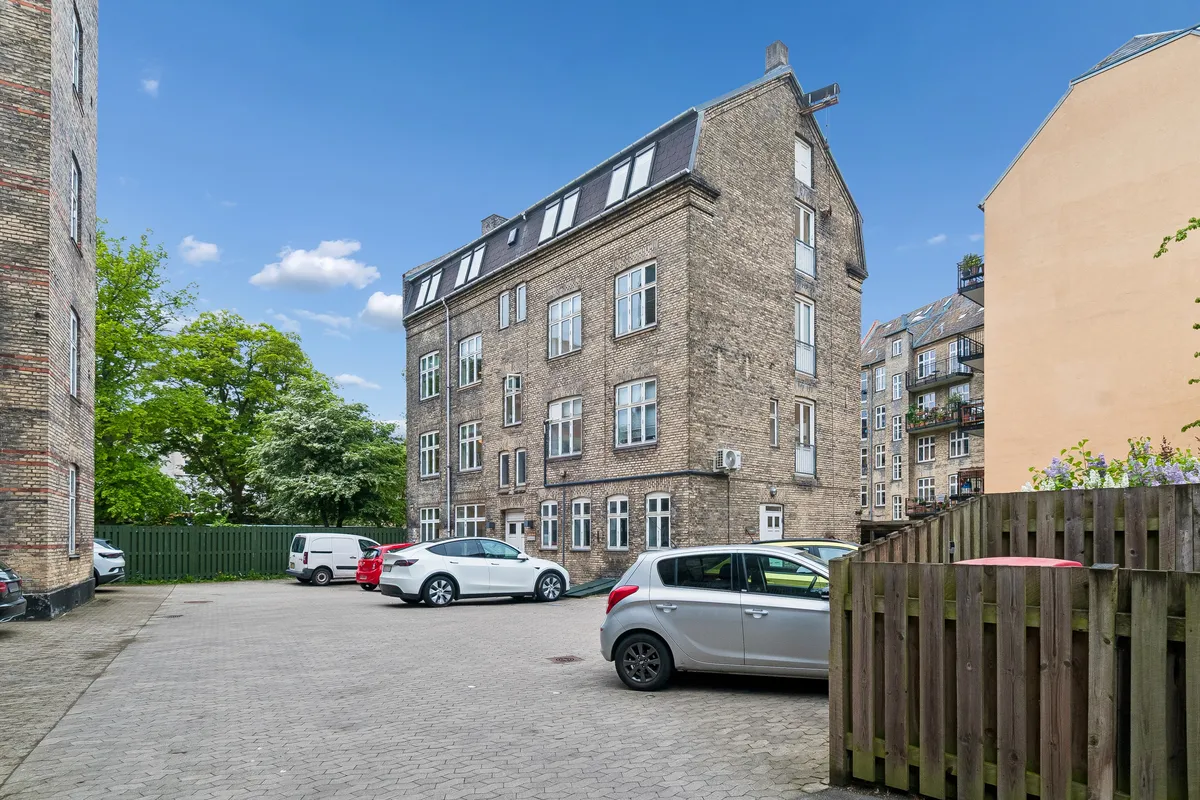 Flere parkerede biler uden for en nyrenoveret bygning med flere lejligheder og terrasser samt lyseruder og plantning i baggården