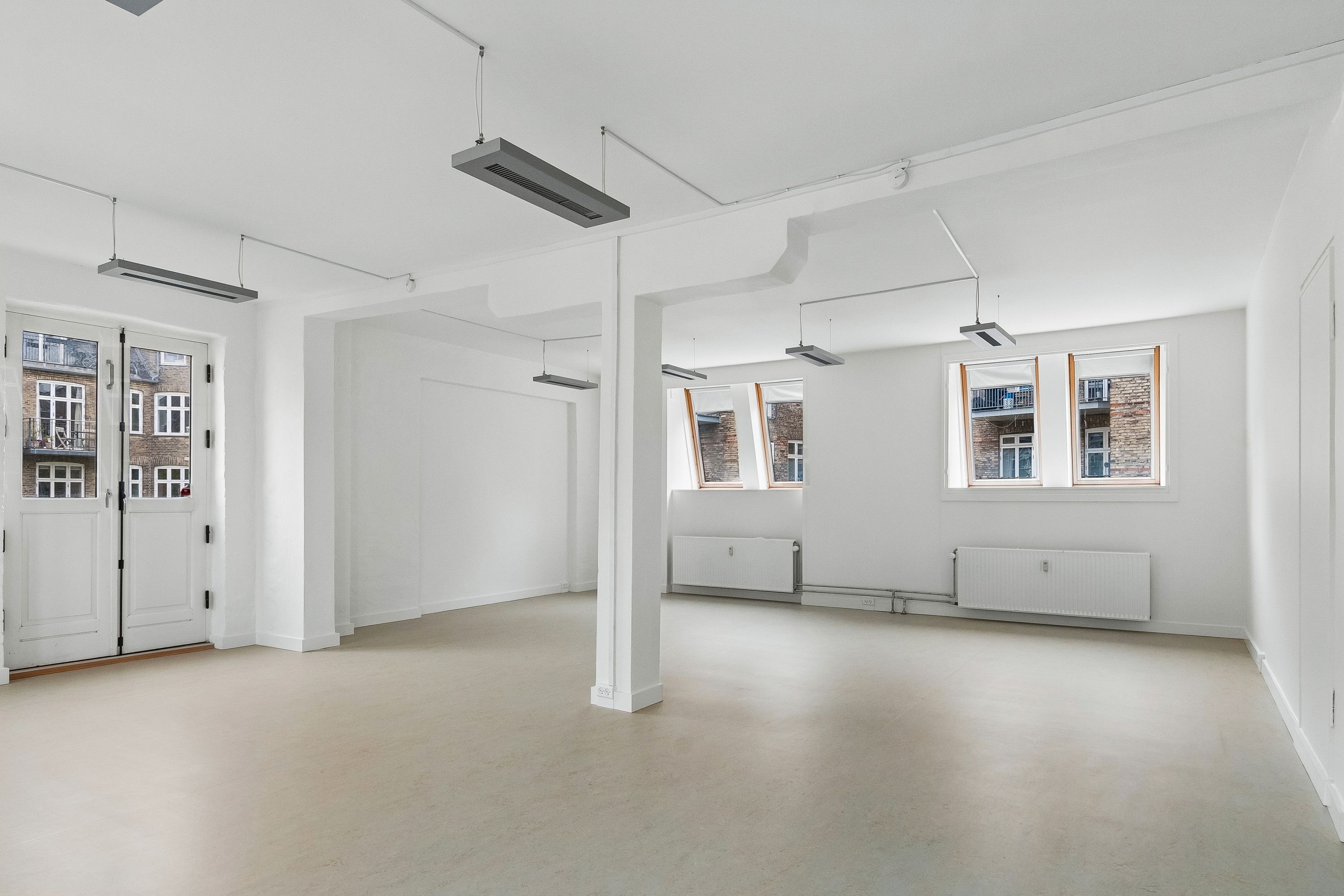 Lyst kontor med hvide vægge og lysegul gulvbelægning. Vinduer med højt gulv og lysegardiner. Moderne radiatorer og loftsmonteret klimaanlæg. Højt gulv til kontorudlejning på Frederiksberg