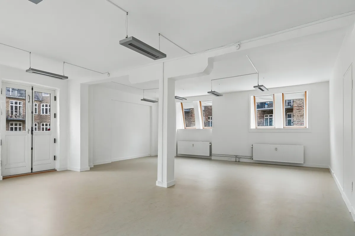 Lyst kontor med hvide vægge og lysegul gulvbelægning. Vinduer med højt gulv og lysegardiner. Moderne radiatorer og loftsmonteret klimaanlæg. Højt gulv til kontorudlejning på Frederiksberg
