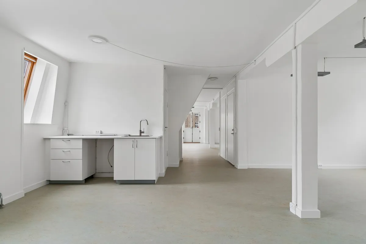 Lyst og rummeligt kontor med hvidt loft, hvide vægge, gulv og møbler samt lyse loftsvinduer og moderne, hvidt køkken