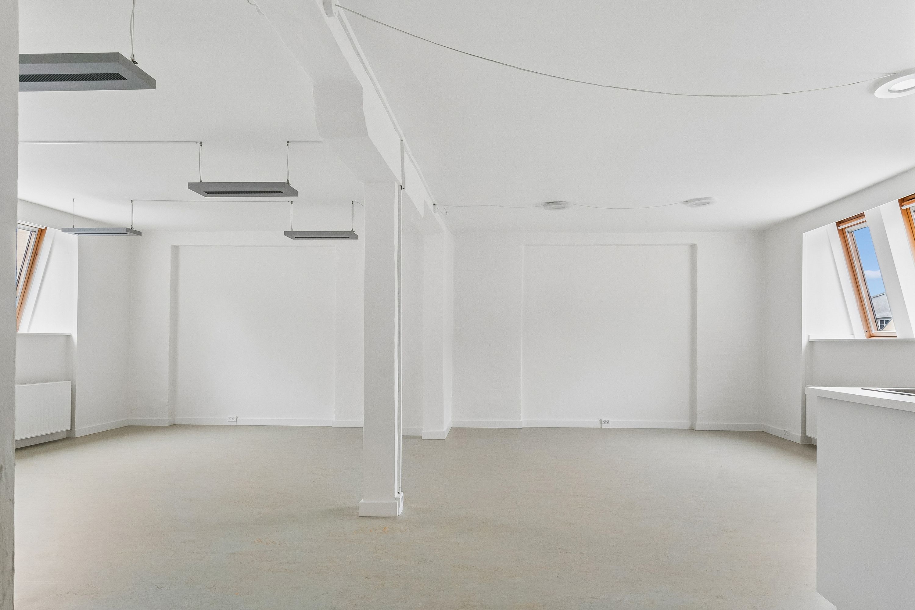 Lyst og rummeligt kontor med hvidt loft og vægge. Store vinduer med trærammer og lysegardiner. Højloftede og højt kvalificeret kontorfællesskab.