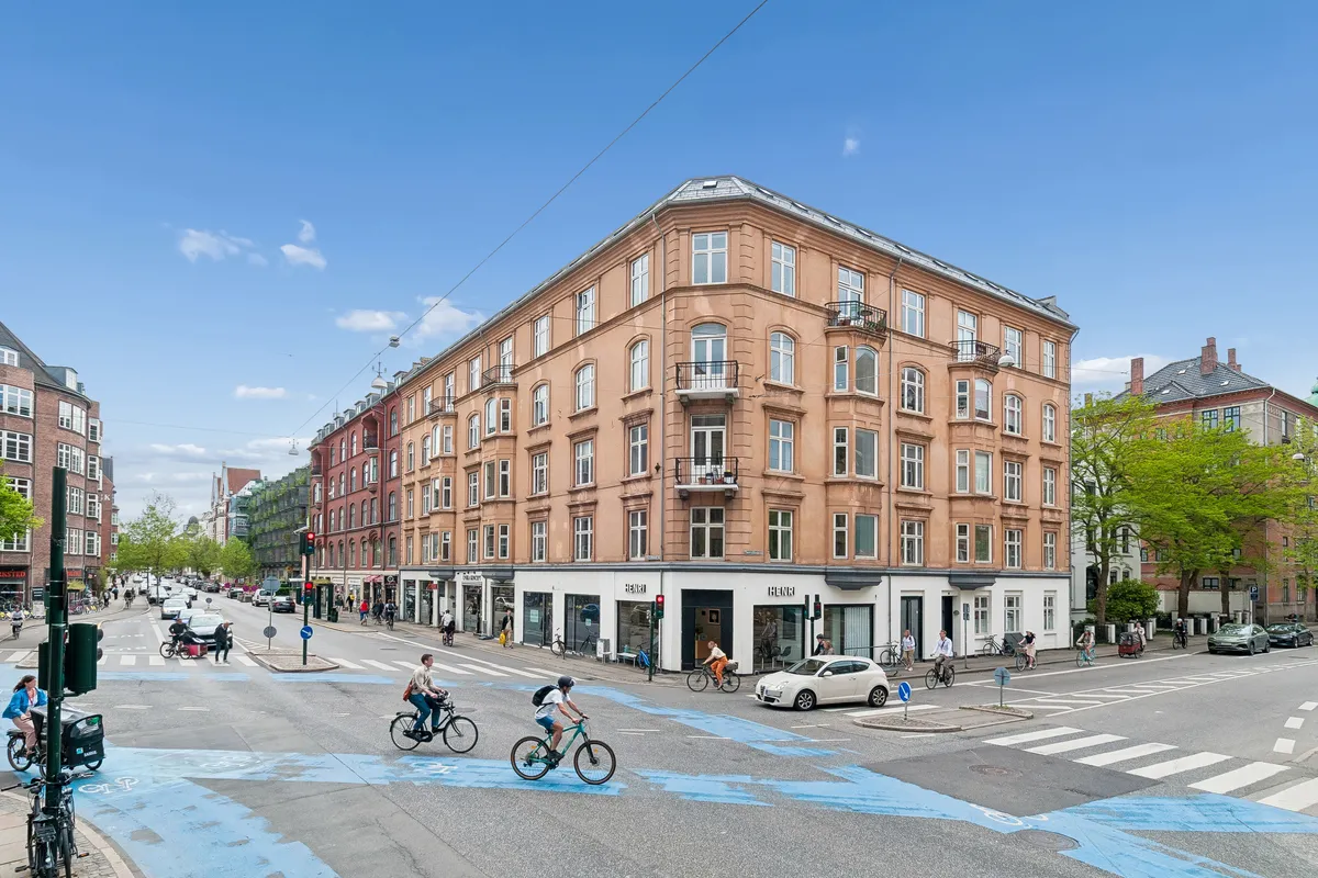 Flere cyklister kører i gaden, og en mand kører på en cykel i den blå farvelagte cykelsti. Bygningen er en stor lejlighedsbygning med mange vinduer og balkoner