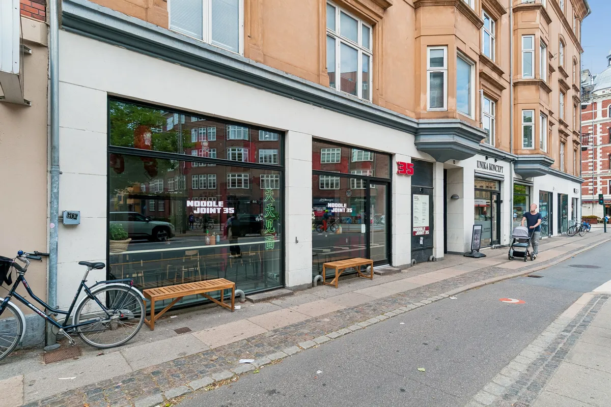 En restaurant med vinduer og teksten Noodle Joint 35 i glasset. To bænke uden for døren og en mand med barnevogn går på fortovet.