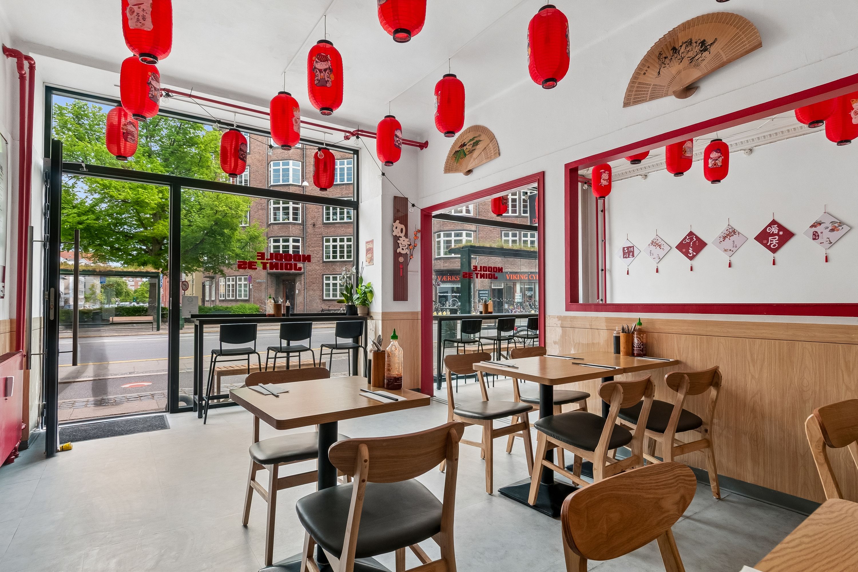 Indvendigt i en restaurant med røde lanterner, lyse træborde og stole, der vender mod en udsigt til gaden med bygninger og en stor træ