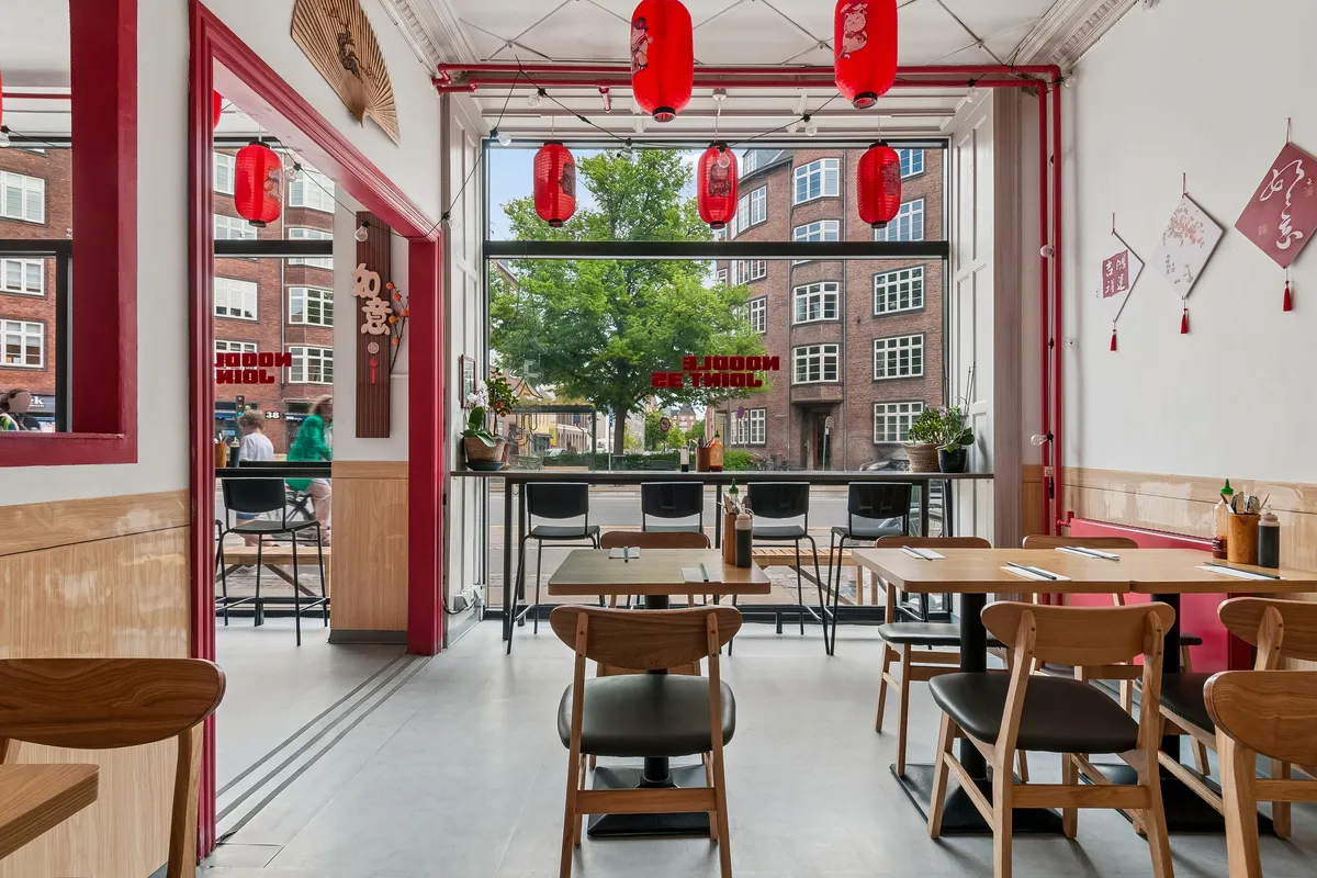 Indvendigt billede af en elegant restaurant med røde lanterner hængende og kinesiske tegn på væggen. Bord og stole er ordnet, og der er plantepotter og lyse vinduer i baggrunden