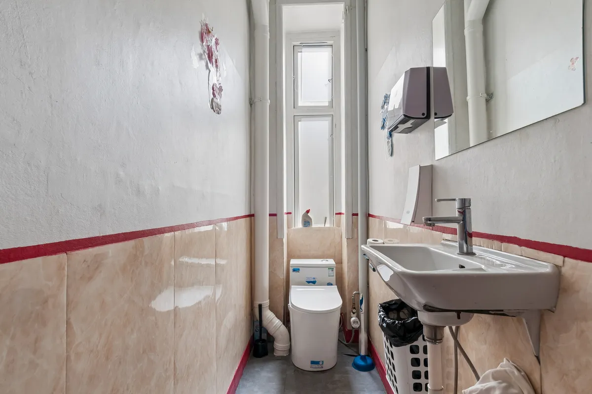 Et moderne toilet i en restaurant med en hvid toilet, et hvidt vask og en spejl på væggen. Der er toiletpapir på væggen, og der er en skraldespand udenfor.