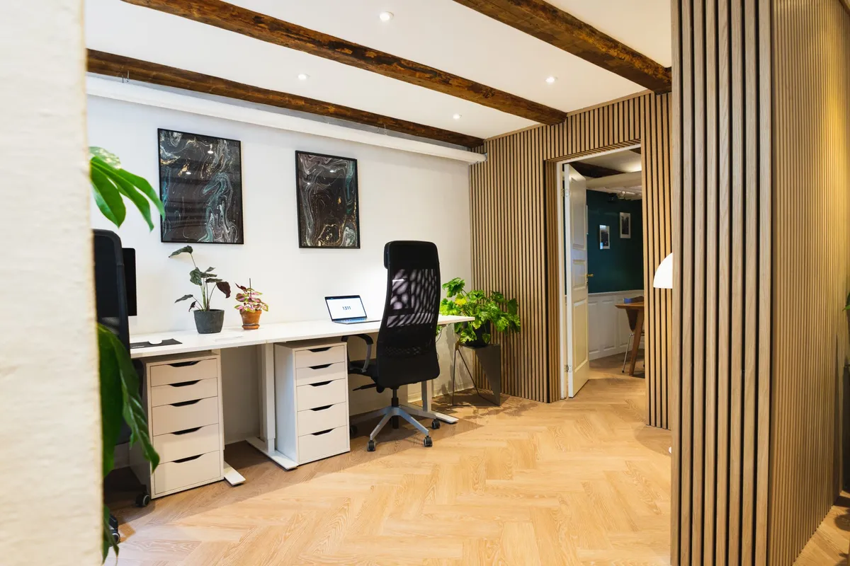 Kontor med højt til loftet, store vinduer og parketgulve. Kontorbord med 2 stole, 2 plantepot og 2 billeder på væggen
