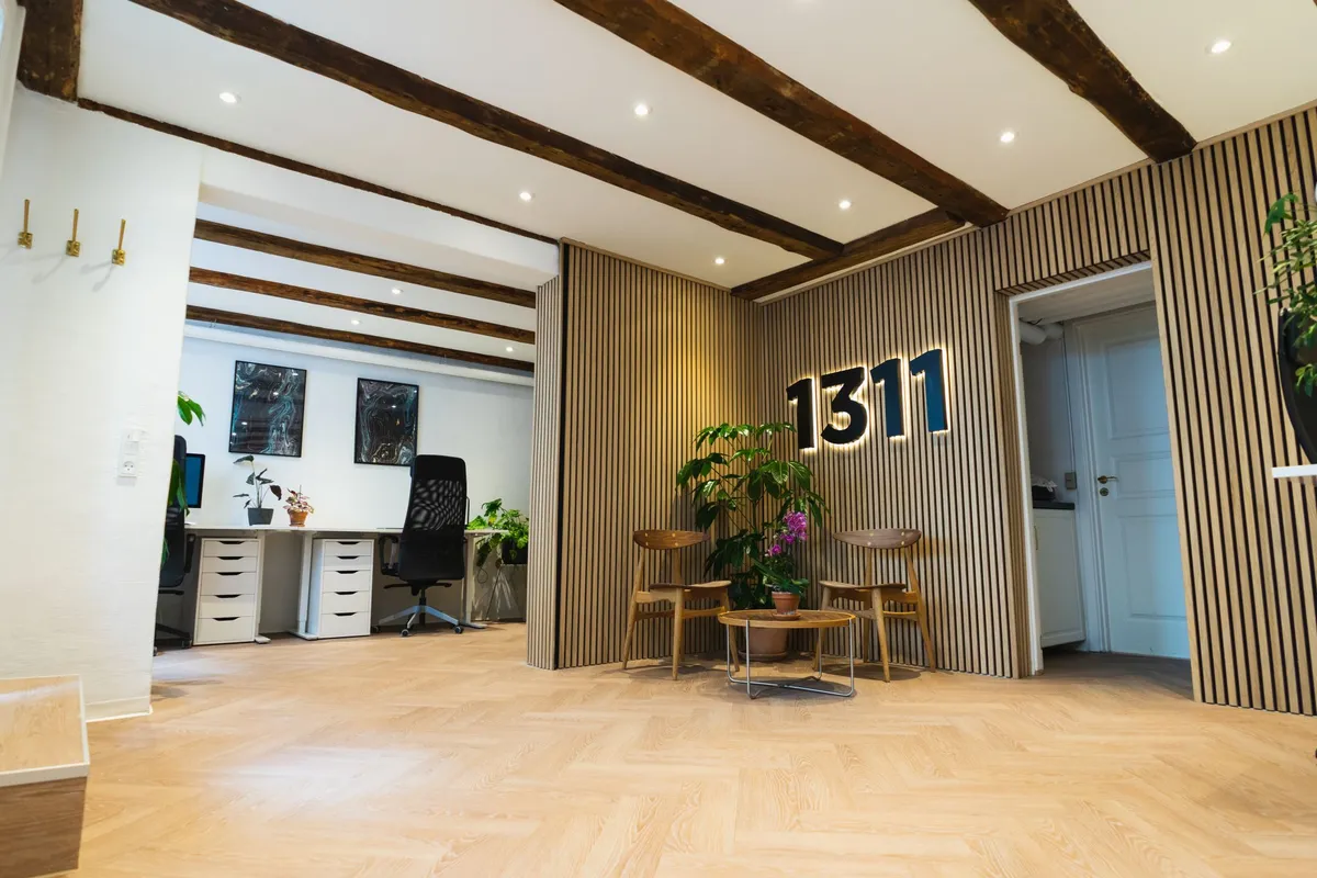 Kontor med moderne møbler og plantepoter på gulvet. 1311 på væggen og en stor plante på siden. Lyse loftsbjælker og møbler i lyse farver