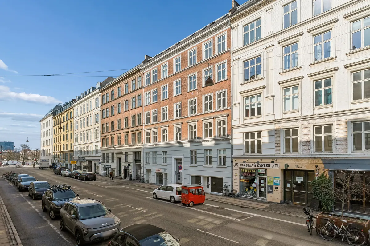 Gade med parkerede biler og gående mennesker foran høje bygninger med mange vinduer og kælderlejligheder