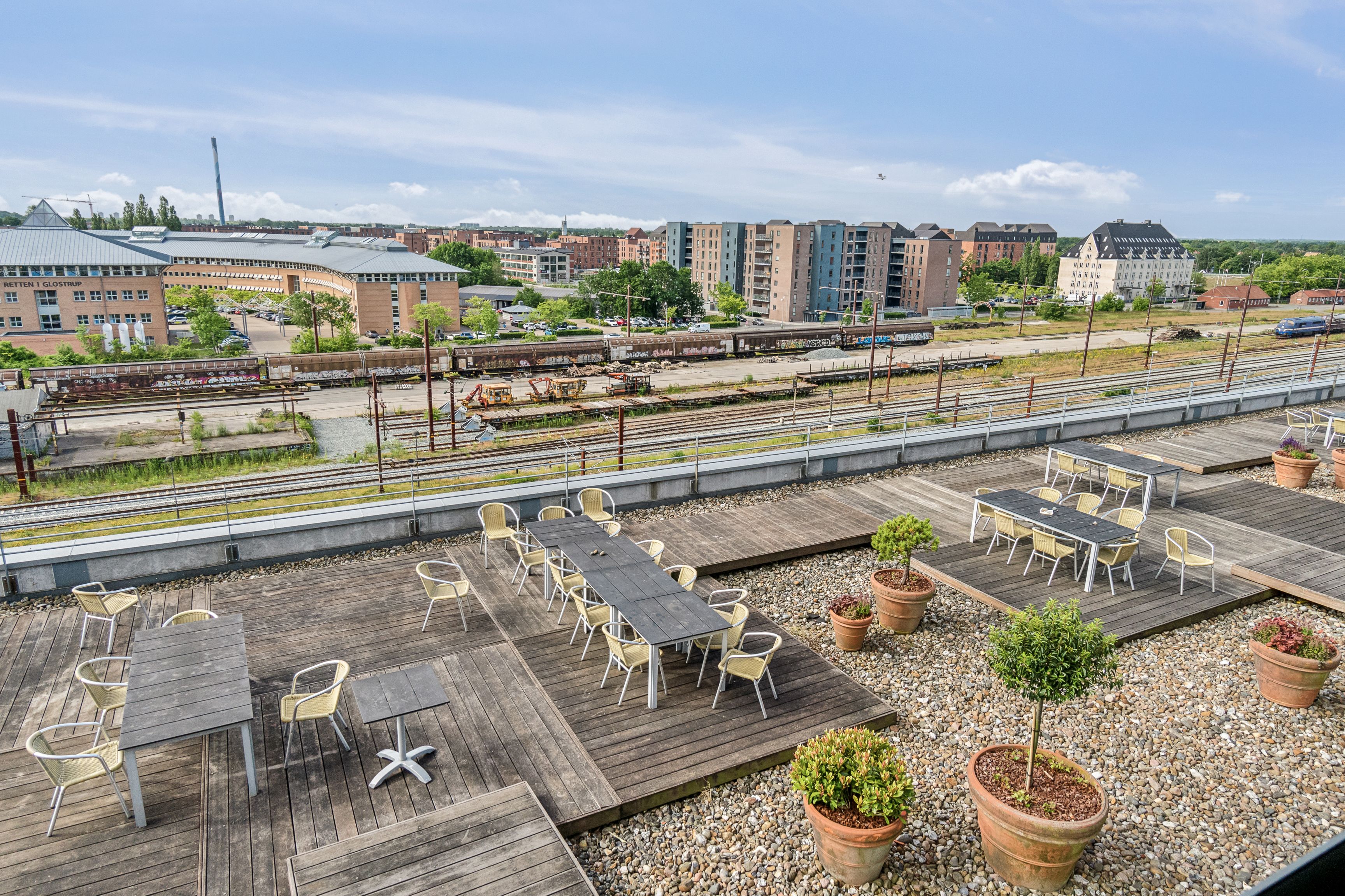 Tagterrasse med smukke udsigt til spor og bygninger i Glostrup Station, forskellige træborde, hvide stole og potteplanter